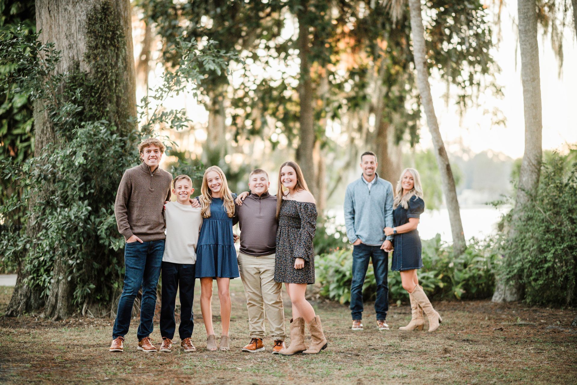 Group of six standing outdoors under trees, smiling in a park at sunset.