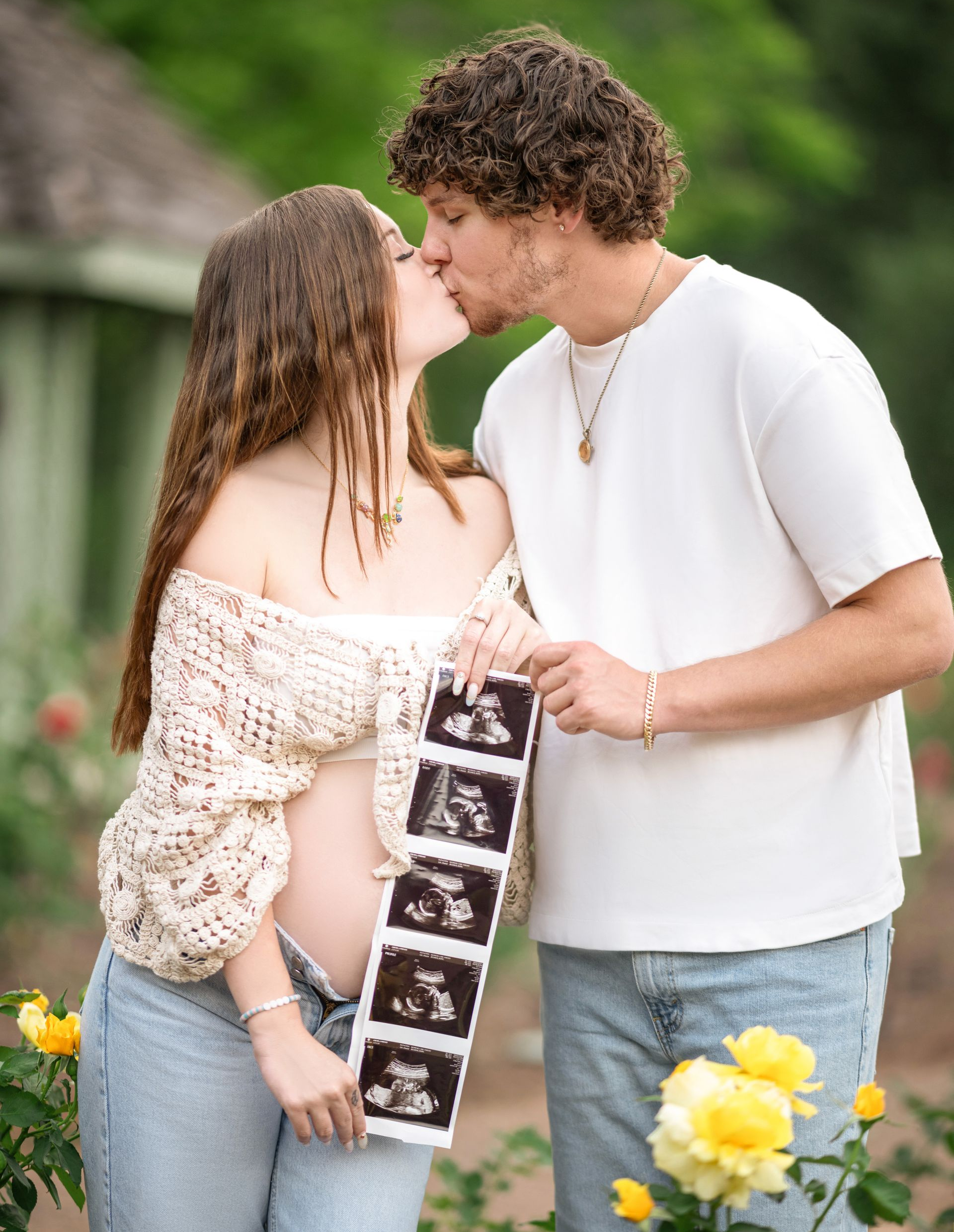 Pregnant couple kissing, holding ultrasound images in a garden with yellow flowers.
