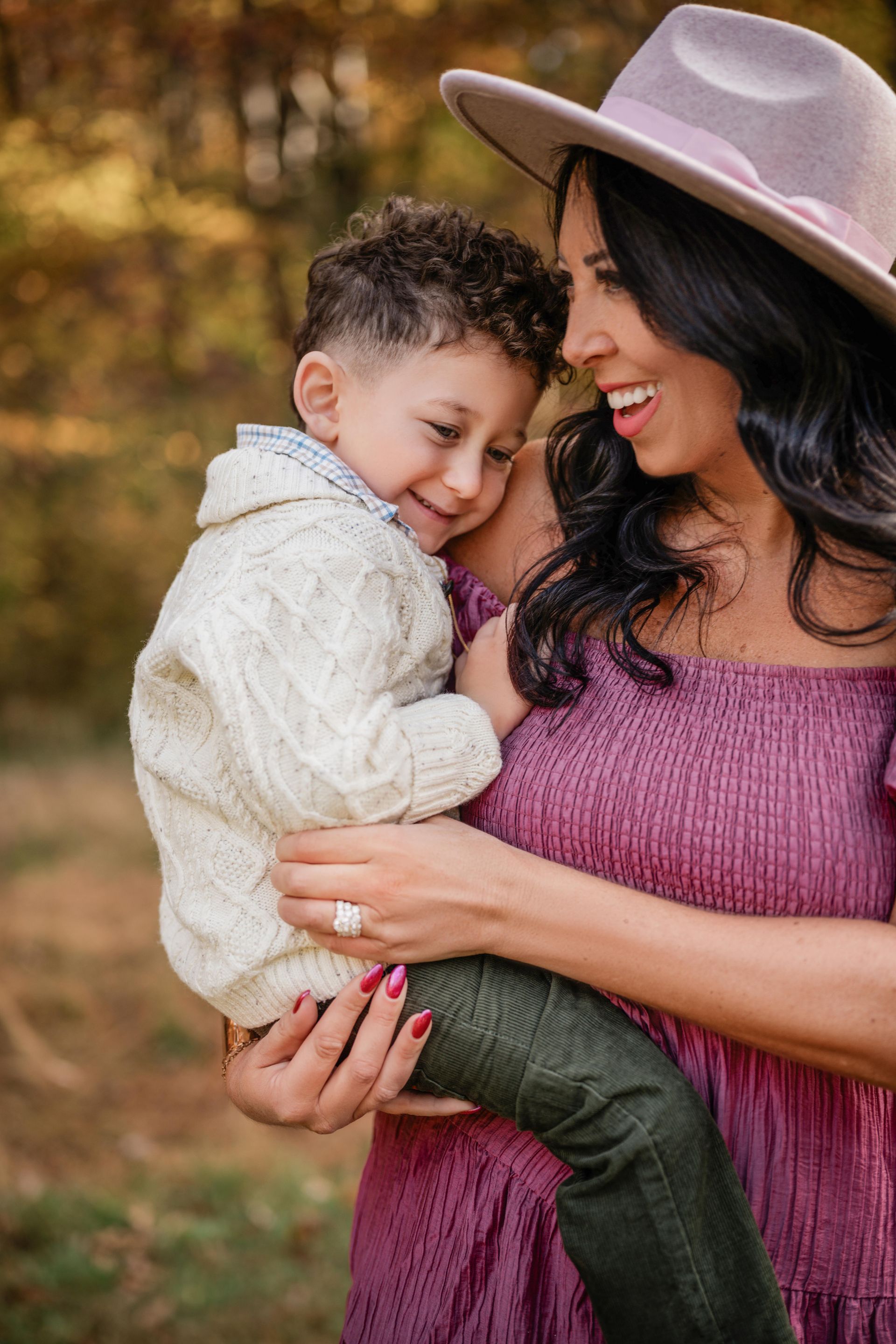 Woman in a mauve dress and hat holding a smiling child in a cream sweater outdoors.