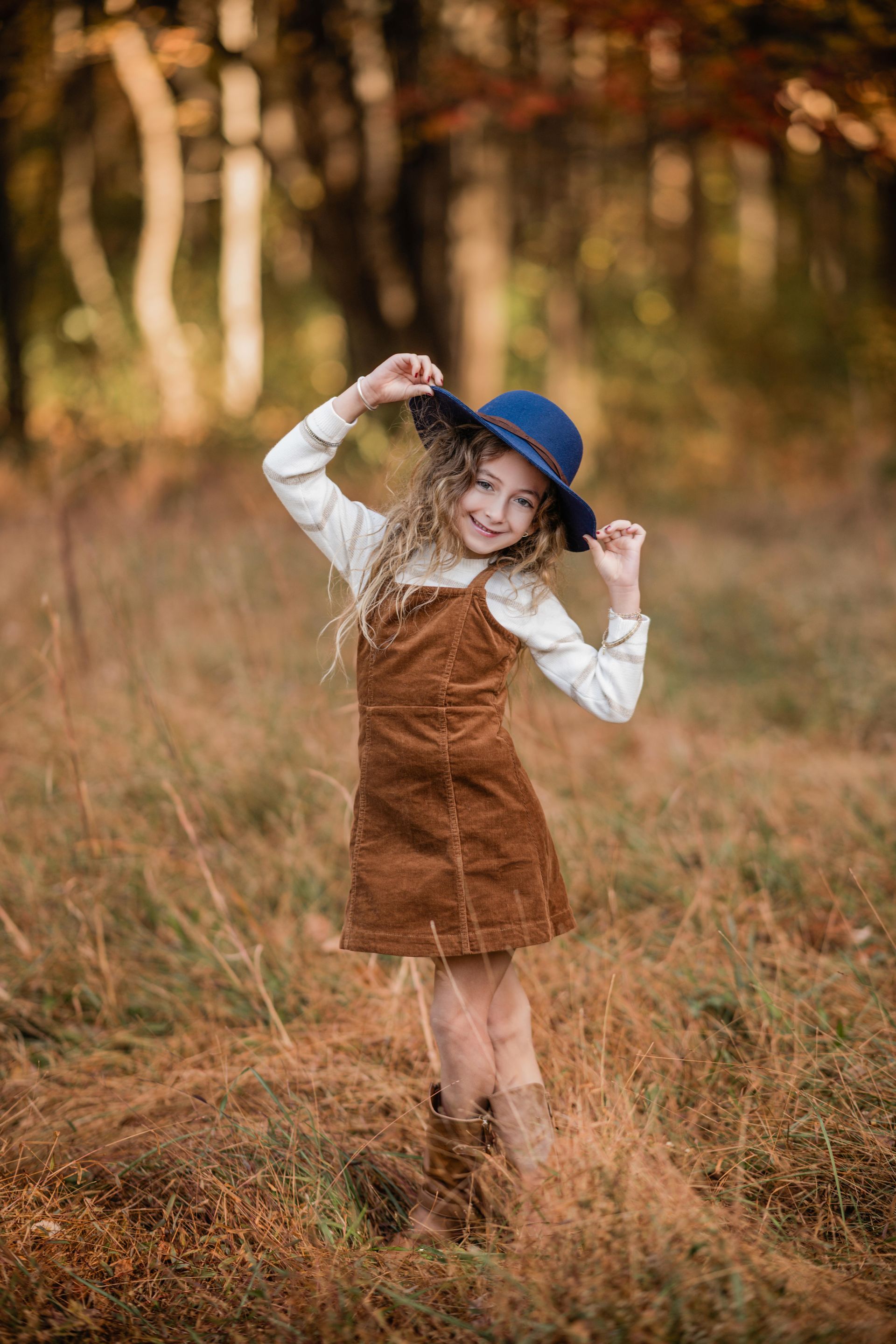 Young child in a blue hat and brown dress standing in tall grass outdoors, smiling and adjusting the hat