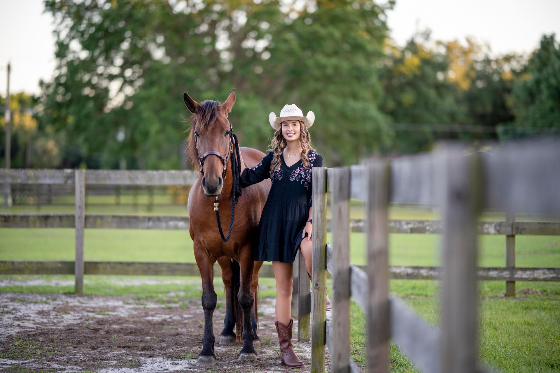 Woman in a white hat stands beside a brown horse in a fenced paddock at sunset.