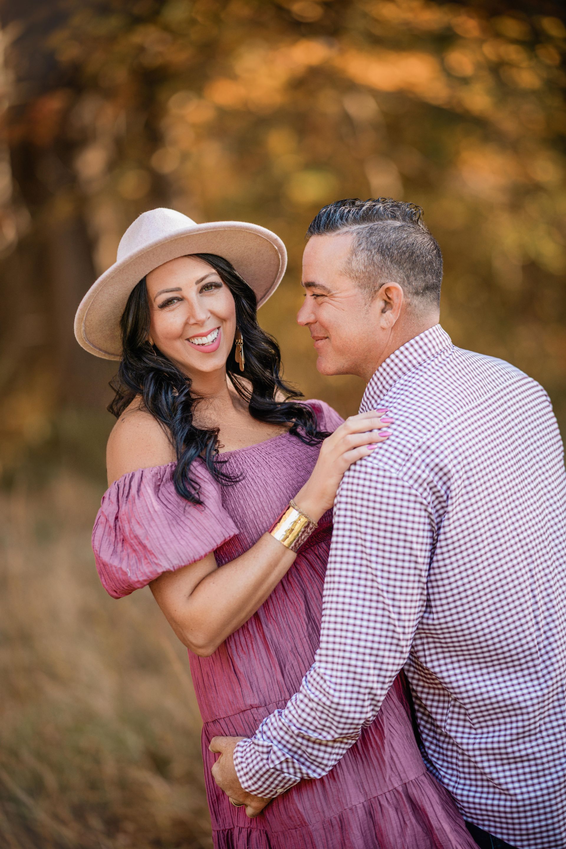 Couple smiling outdoors, the woman in a pink dress and hat leaning on a man in a checkered shirt.