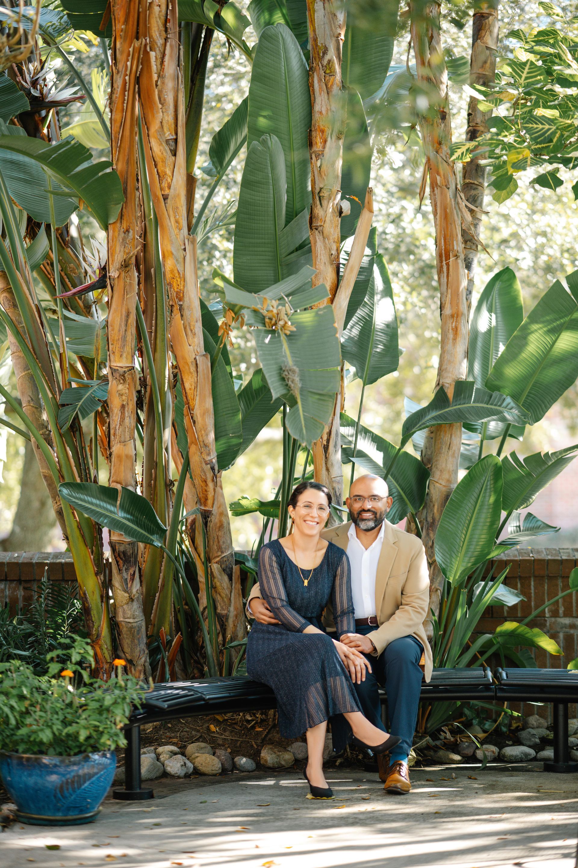 Two people sit on a bench in a lush garden with tall tropical plants and a stone planter.