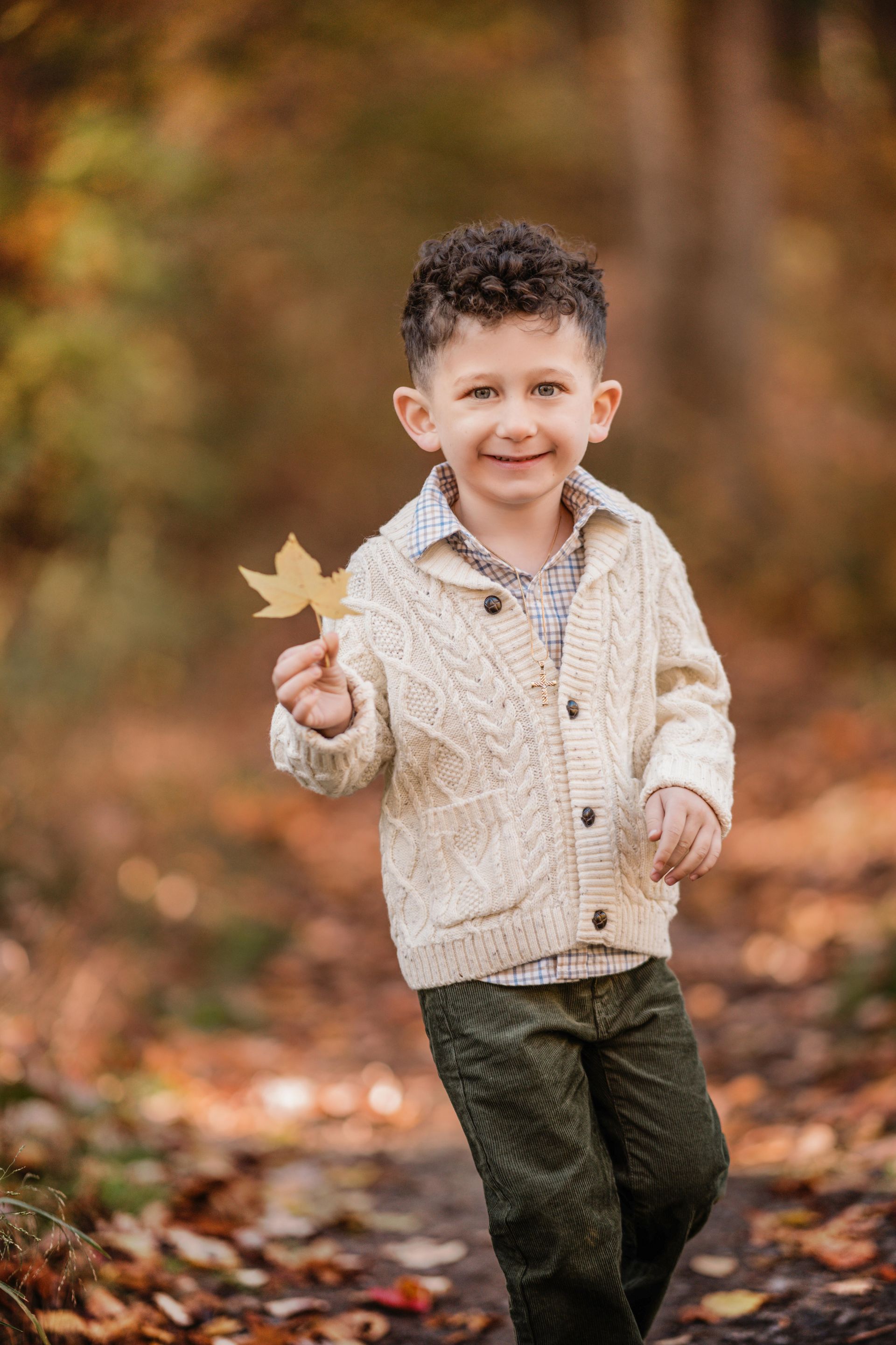 Child walking outdoors on a leafy path, holding up a yellow leaf and smiling.