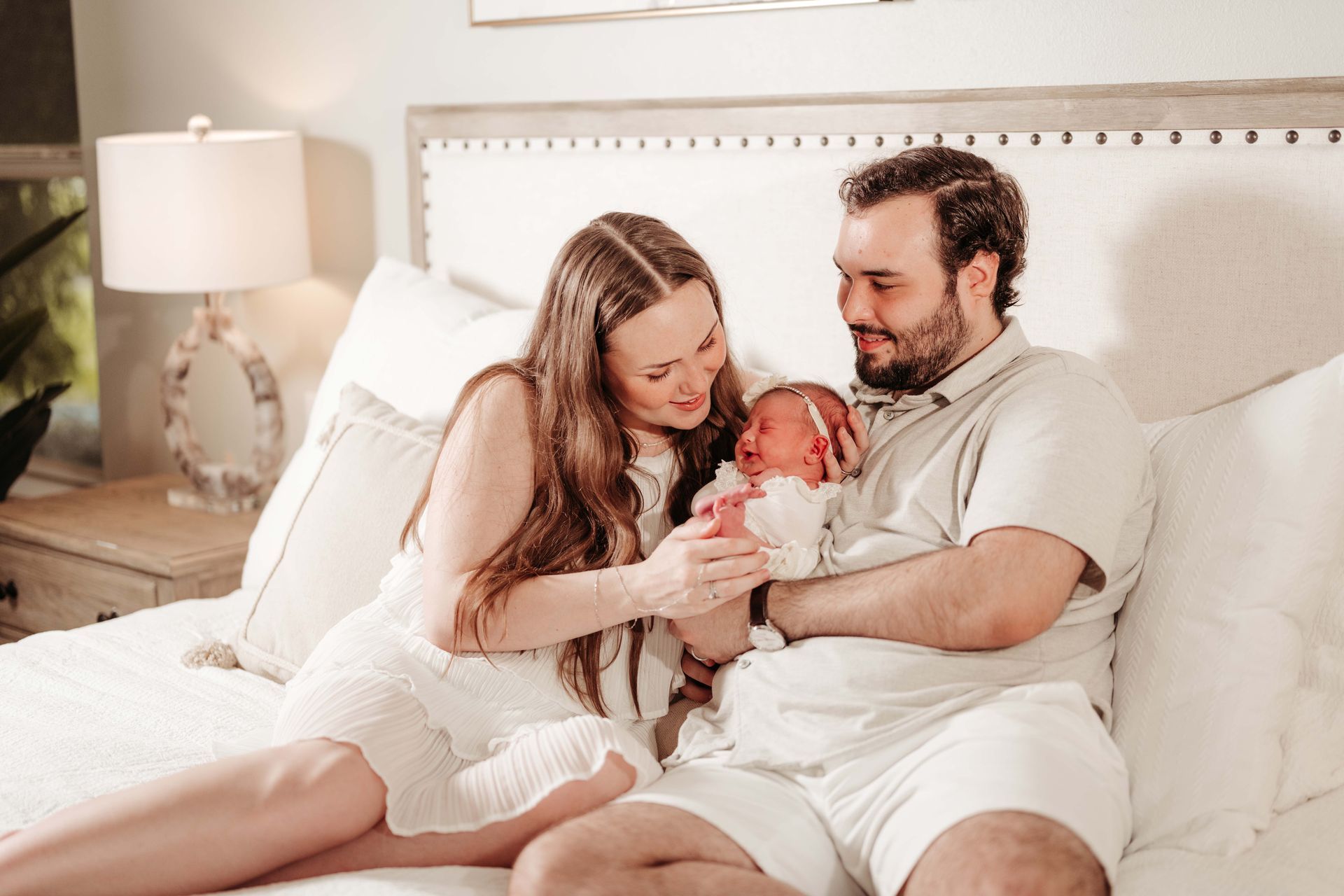 Couple in bed, smiling at newborn baby. White room setting.