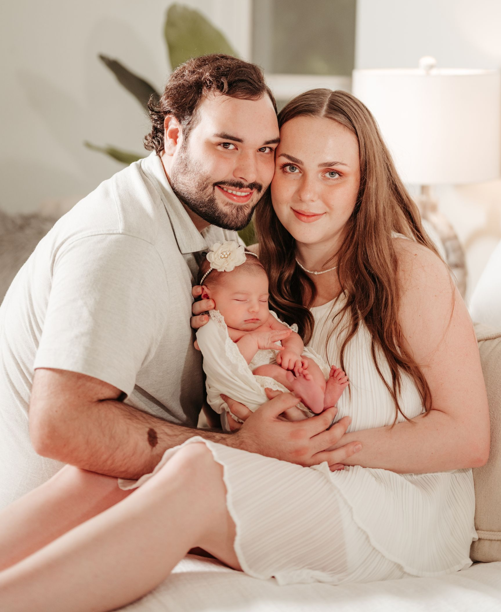 Parents cradling a newborn baby. All wear white clothing, smiling. Soft, well-lit room.