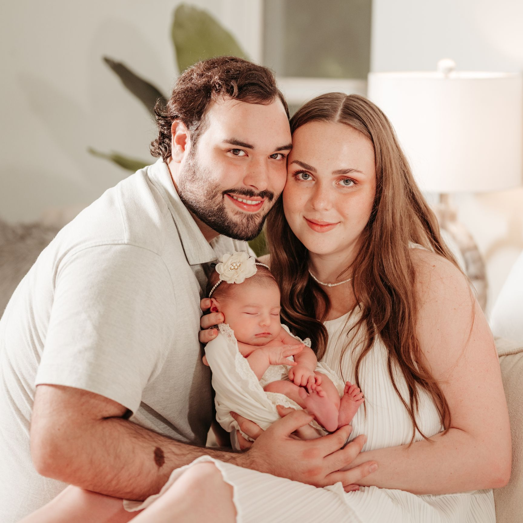 Family of three smiles at camera; parents holding newborn. Indoor setting with soft lighting.