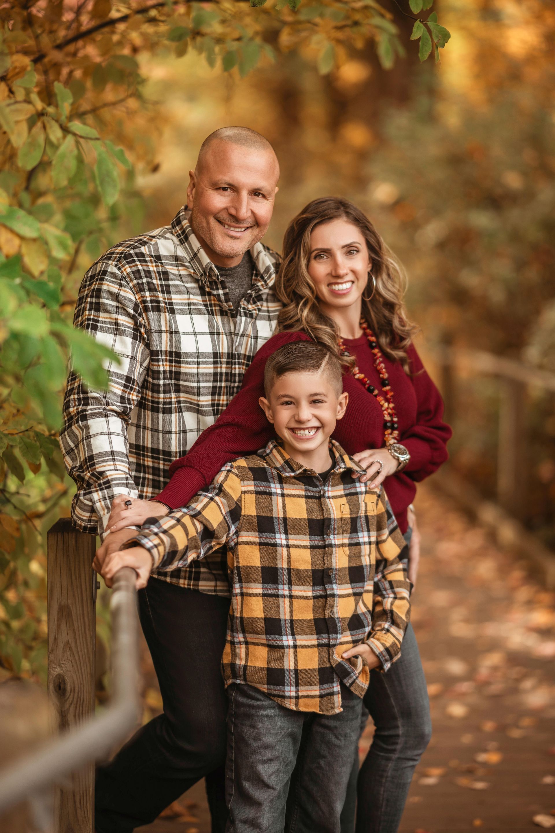 Three people smiling outdoors on an autumn path, leaning on a wooden fence