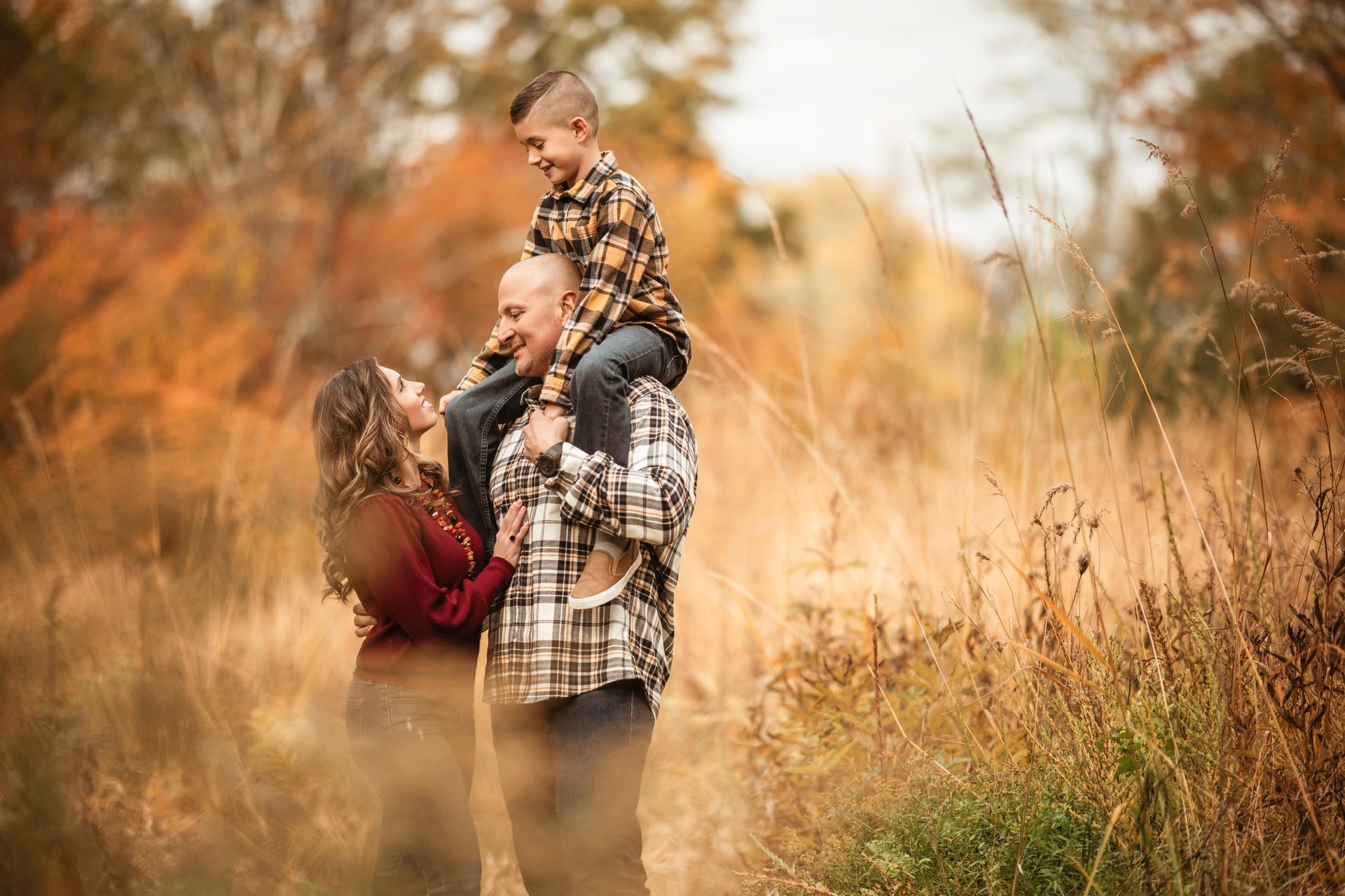Family of three in plaid outdoors on a grassy path, with a child sitting on a man’s shoulders and a woman beside them.