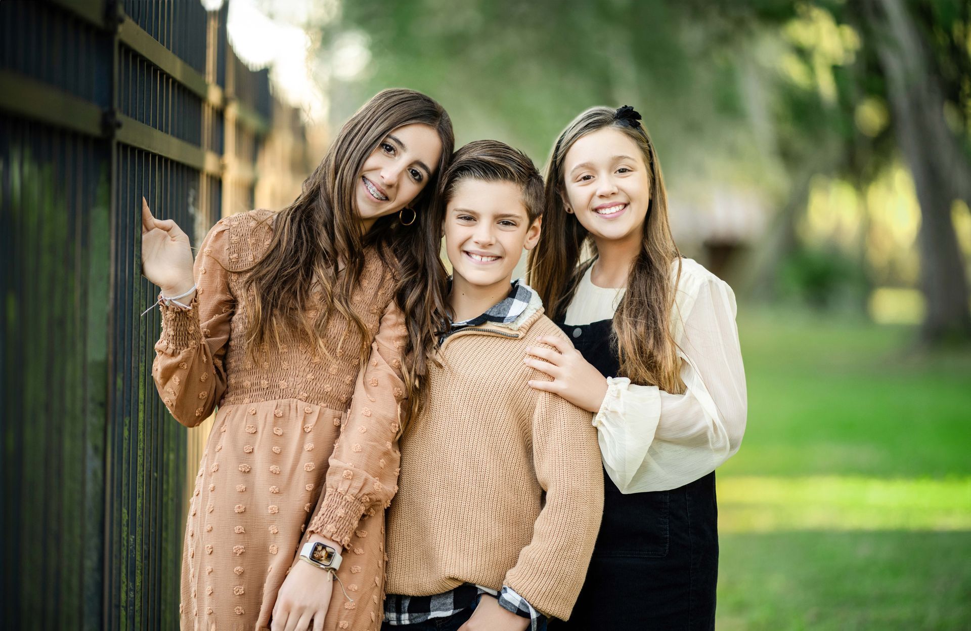 Three smiling friends standing outdoors by a fence, one waving, in warm casual outfits.
