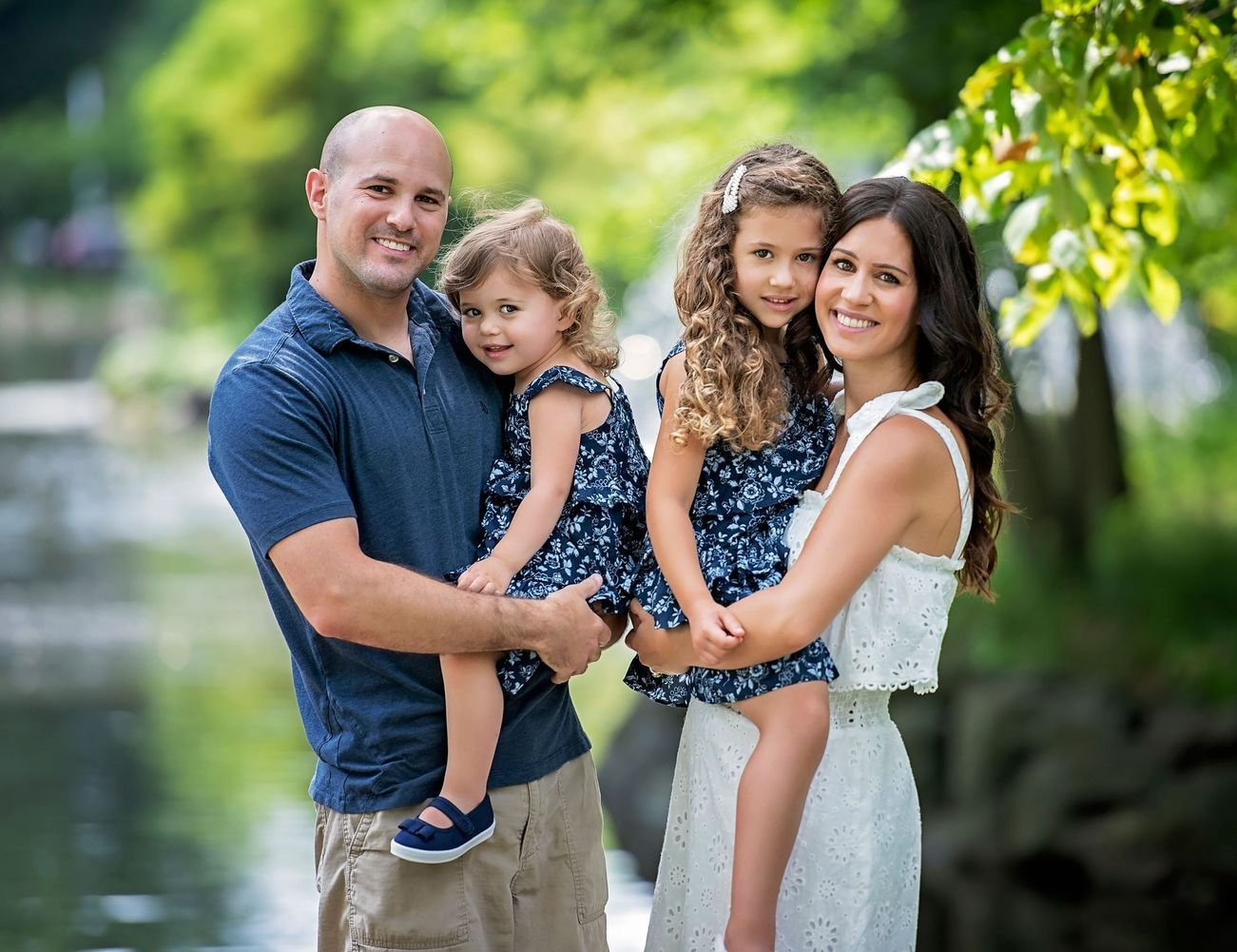 Family of four posing outdoors; man holding daughter, woman holding daughter, all smiling.