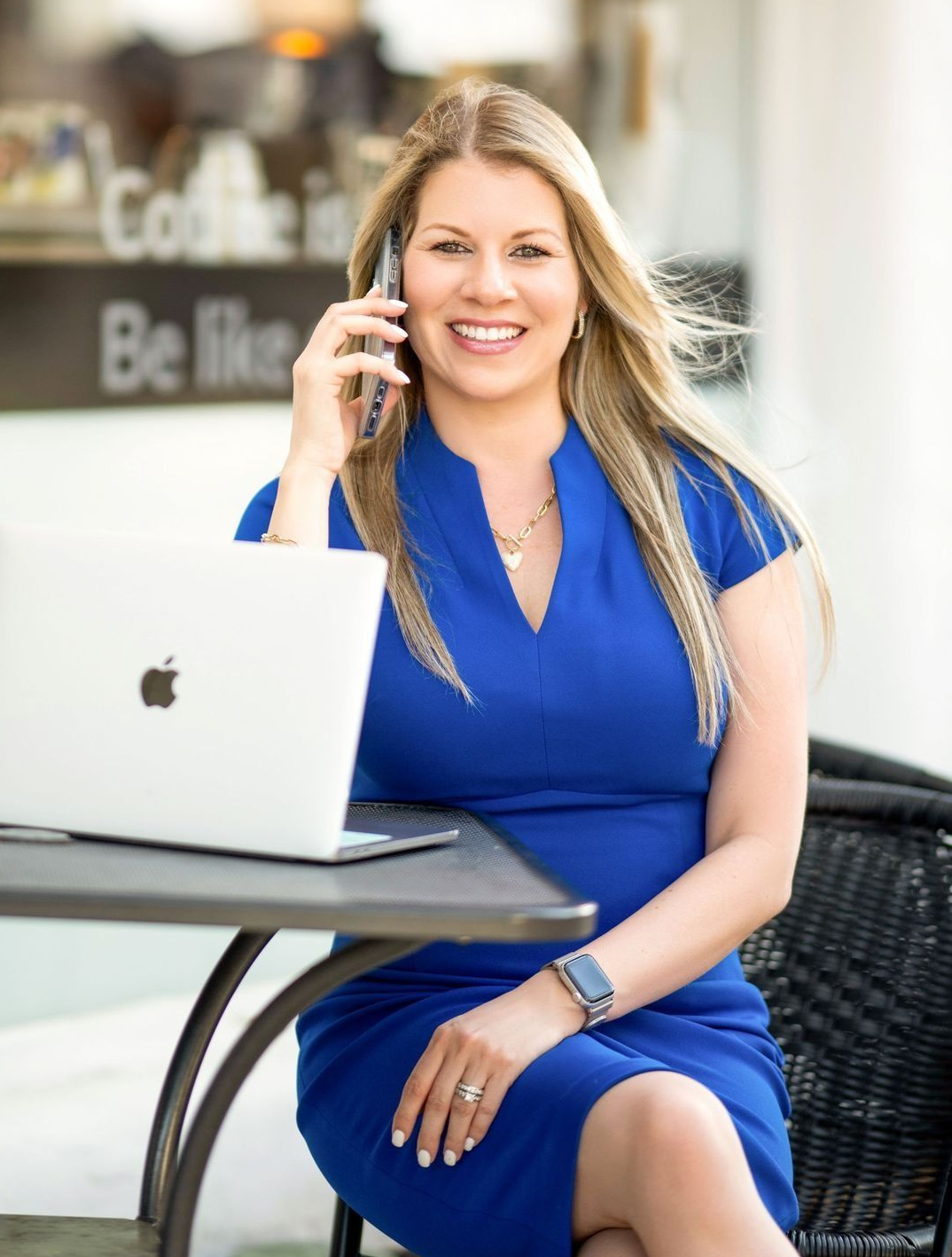 Woman in blue dress smiles, talks on phone, works on laptop at outdoor cafe table.