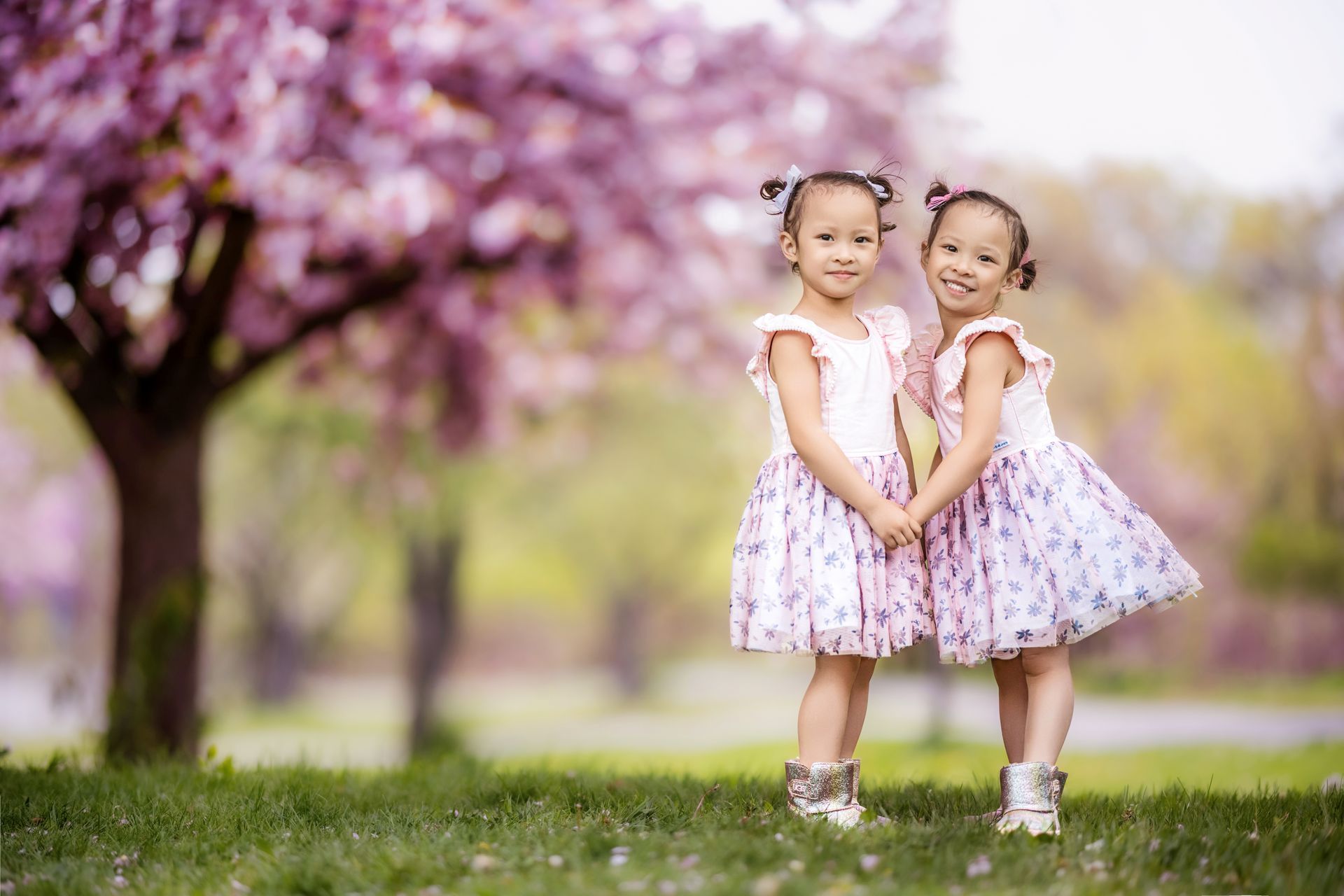 Two children in pink dresses stand on grass beneath blooming trees, smiling and holding hands.