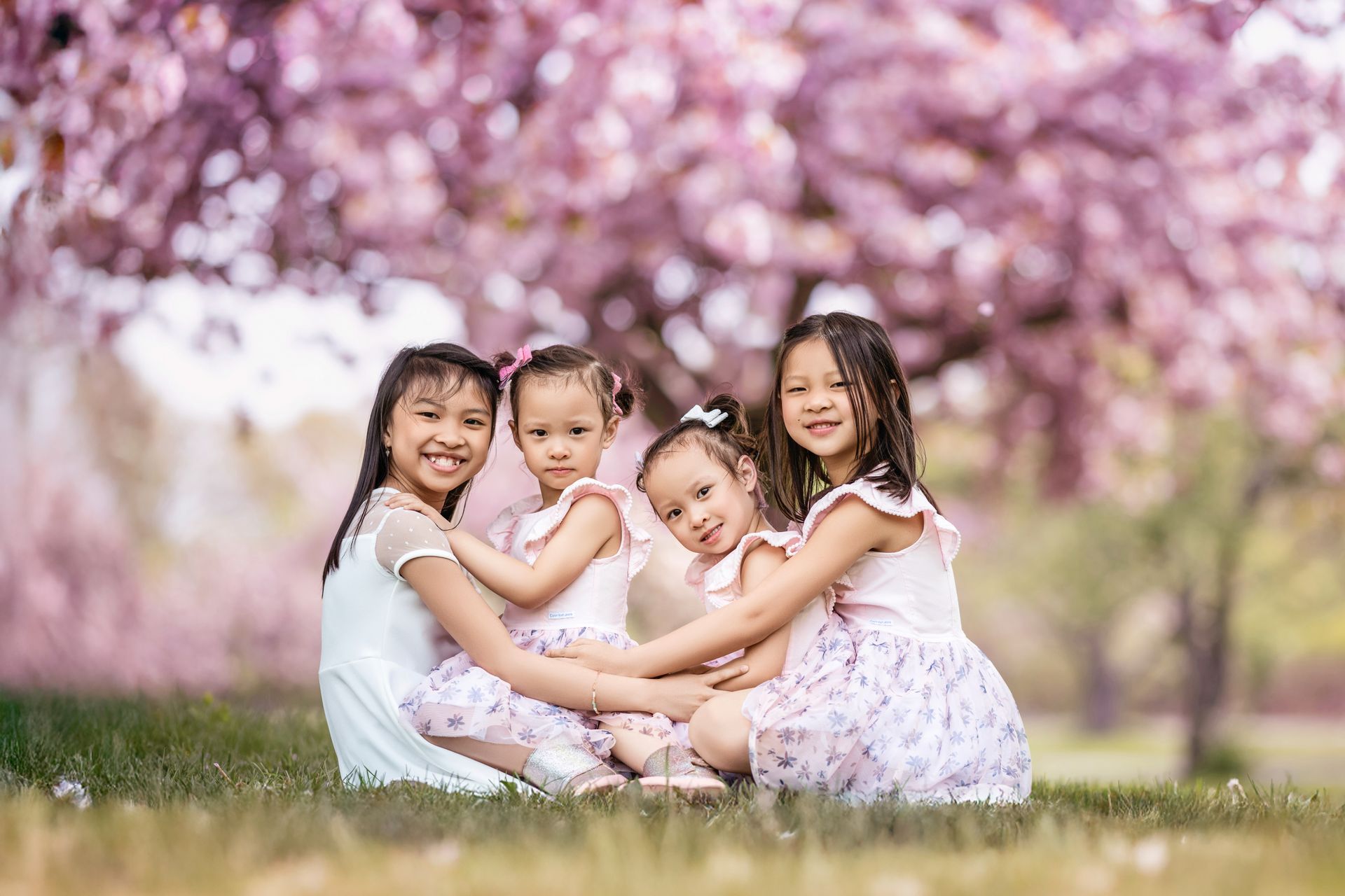 Four children sit on grass beneath pink flowering trees, smiling and hugging.