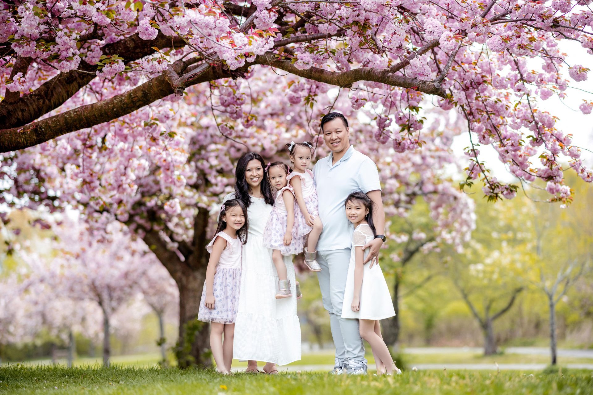Family standing under pink cherry blossoms in a park, smiling and posing together.