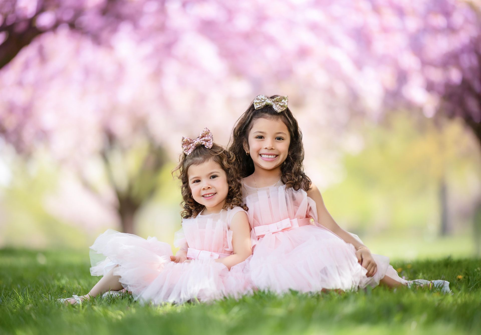 Two children in pink dresses sitting on grass with blooming trees in the background