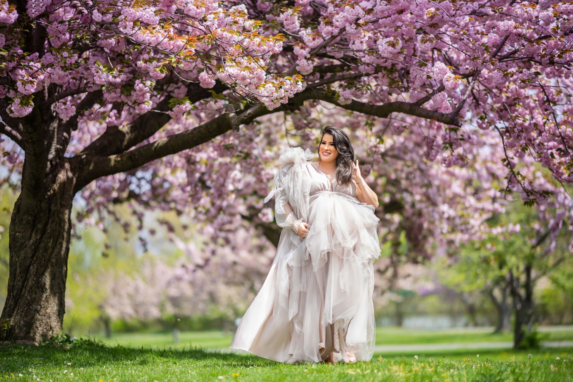 Pregnant woman in a flowing gown under a blossoming cherry tree, holding her stomach.
