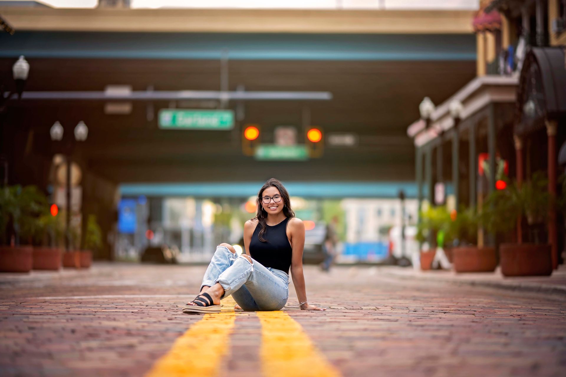 Woman in black top and jeans, sitting on a brick road with yellow lines, in a city setting.