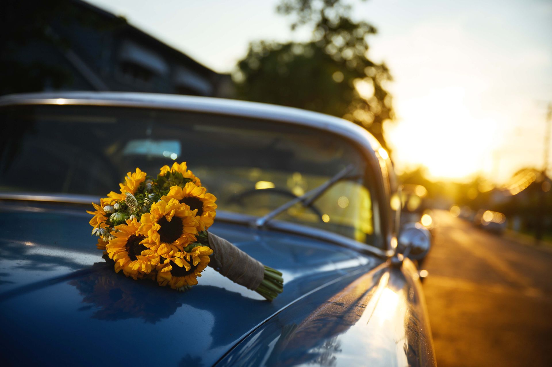 Bouquet of yellow sunflowers on the hood of a classic blue car, with a blurred sunset in the background.