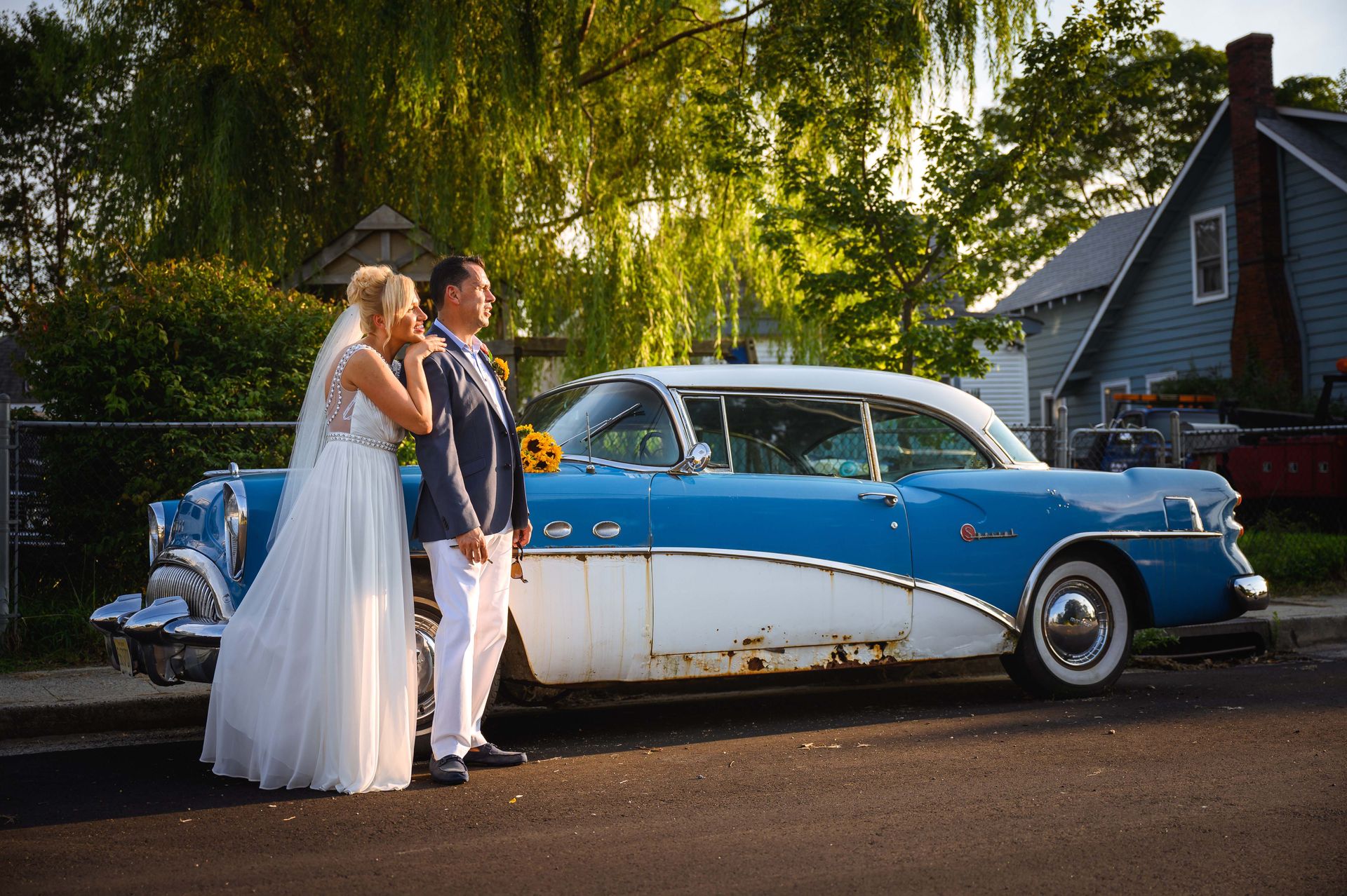 Bride and groom pose beside a vintage blue and white car on a sunny street.