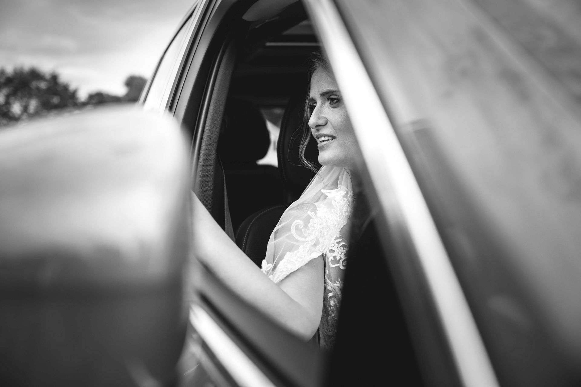 Bride in wedding dress smiles while driving car. Black and white.