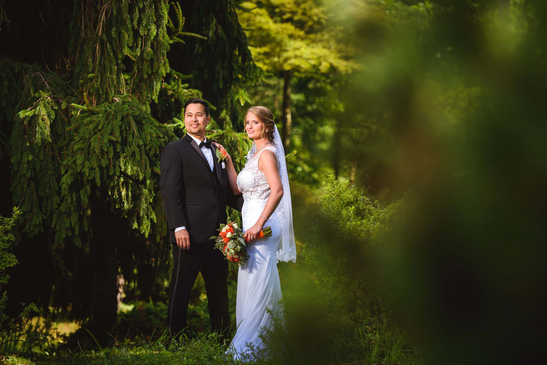 Bride and groom pose in a forest. She wears a white dress, he wears a black tuxedo. Sunlight shines through trees.