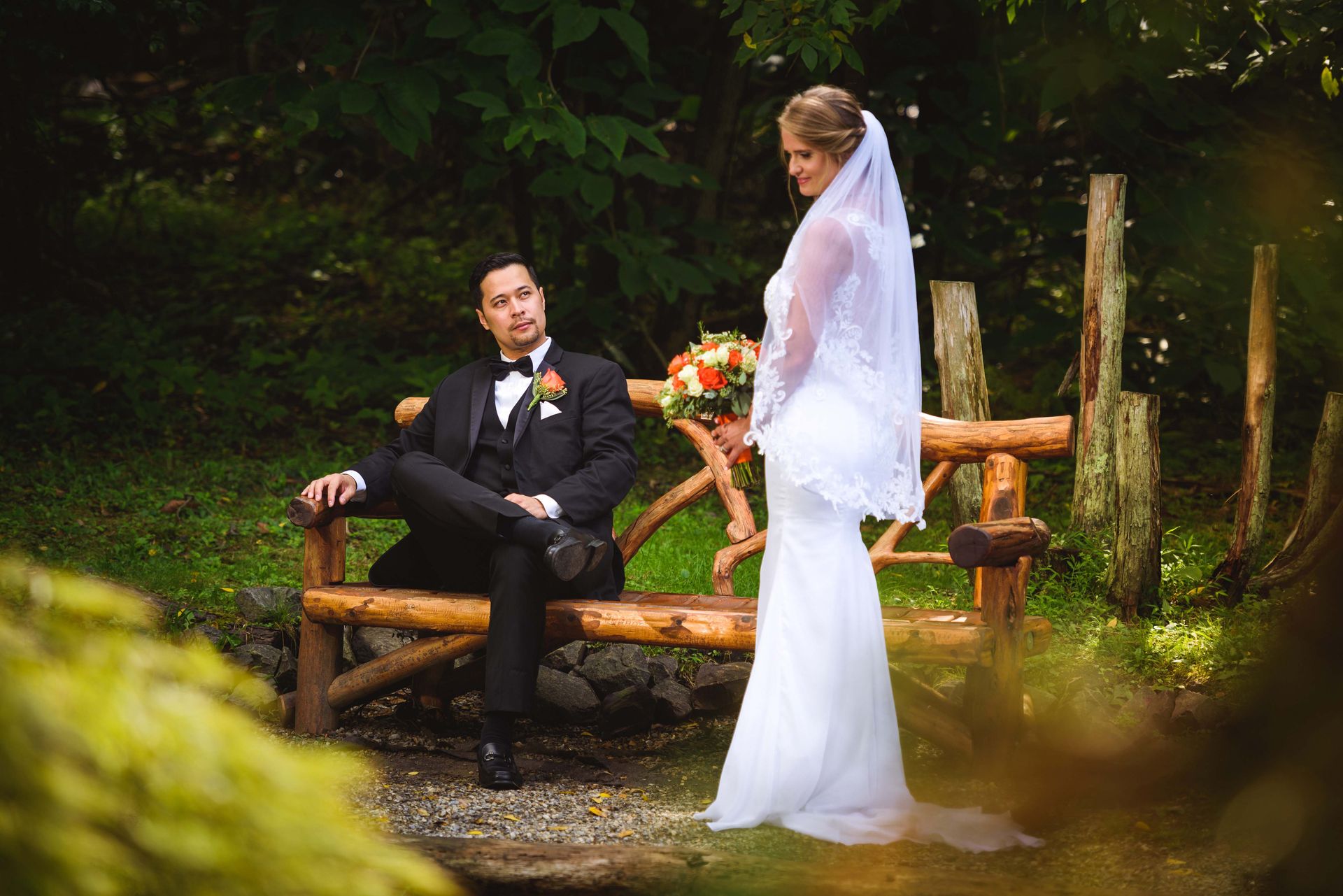 Groom in tuxedo on rustic bench looks at bride in white gown with bouquet and veil in a wooded area.