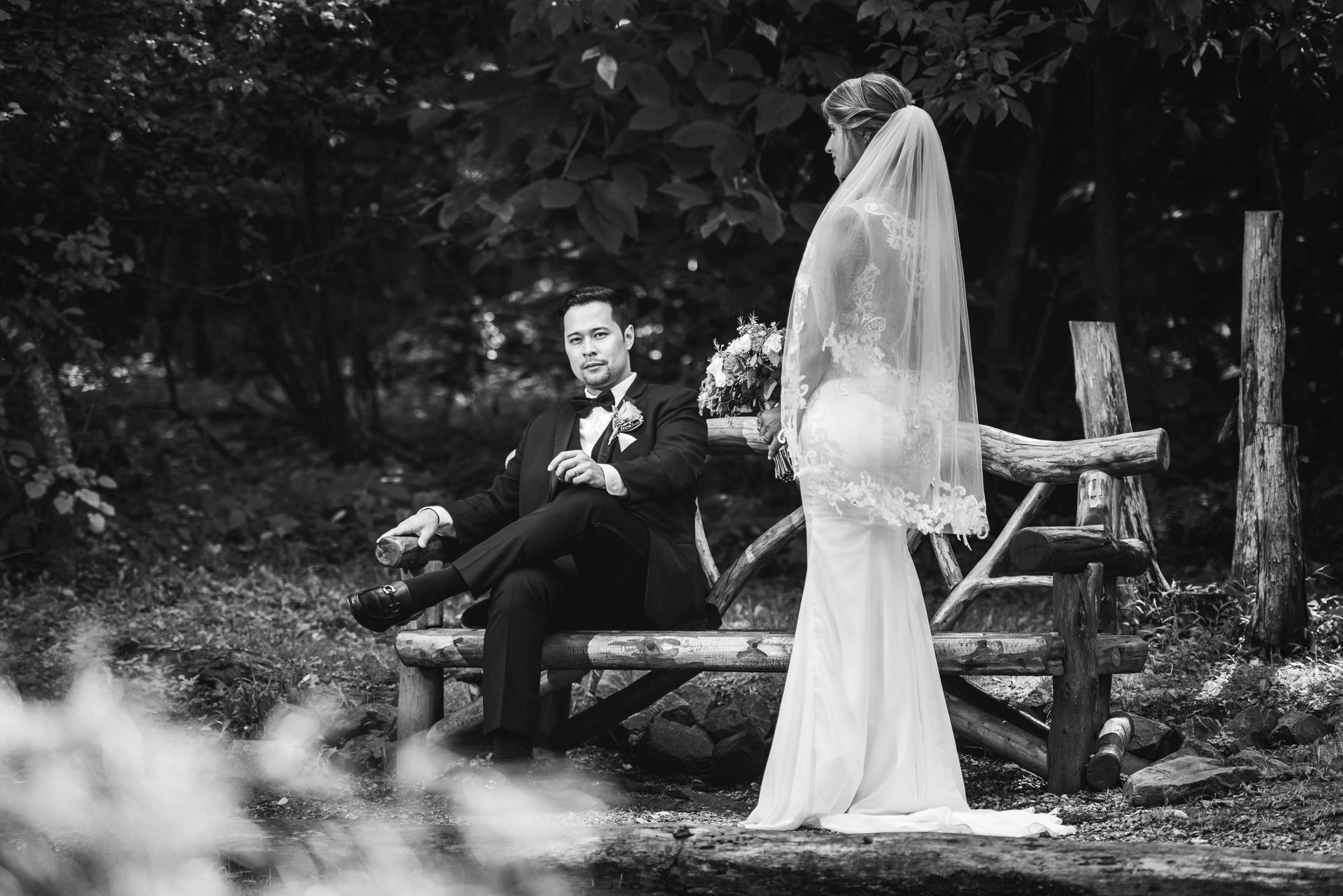 Bride and groom pose outdoors; groom seated on a wooden bench, bride standing next to him in a white dress.