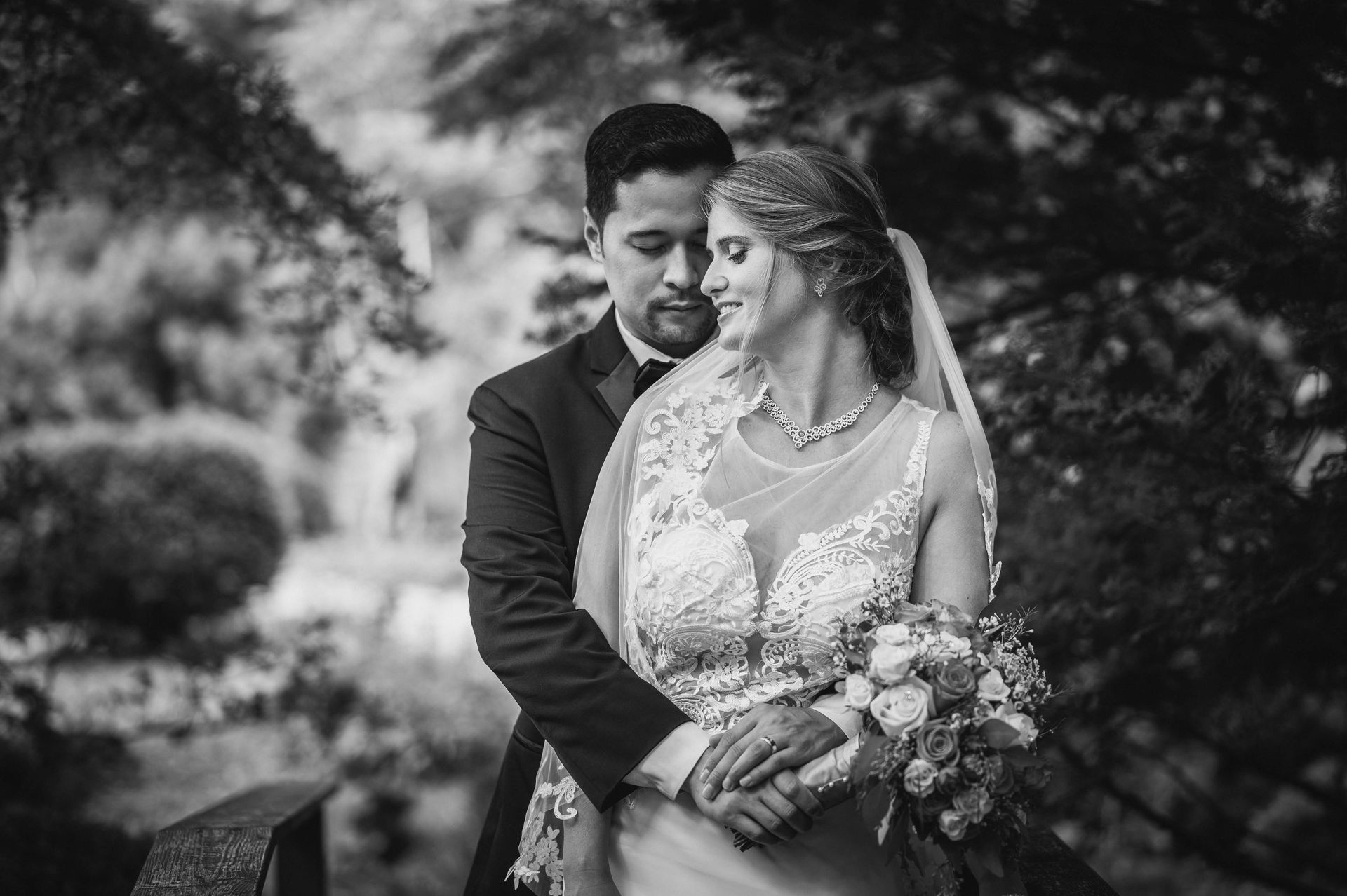 Black and white photo of a newlywed couple embracing outdoors. The bride wears a white gown, the groom a tuxedo.