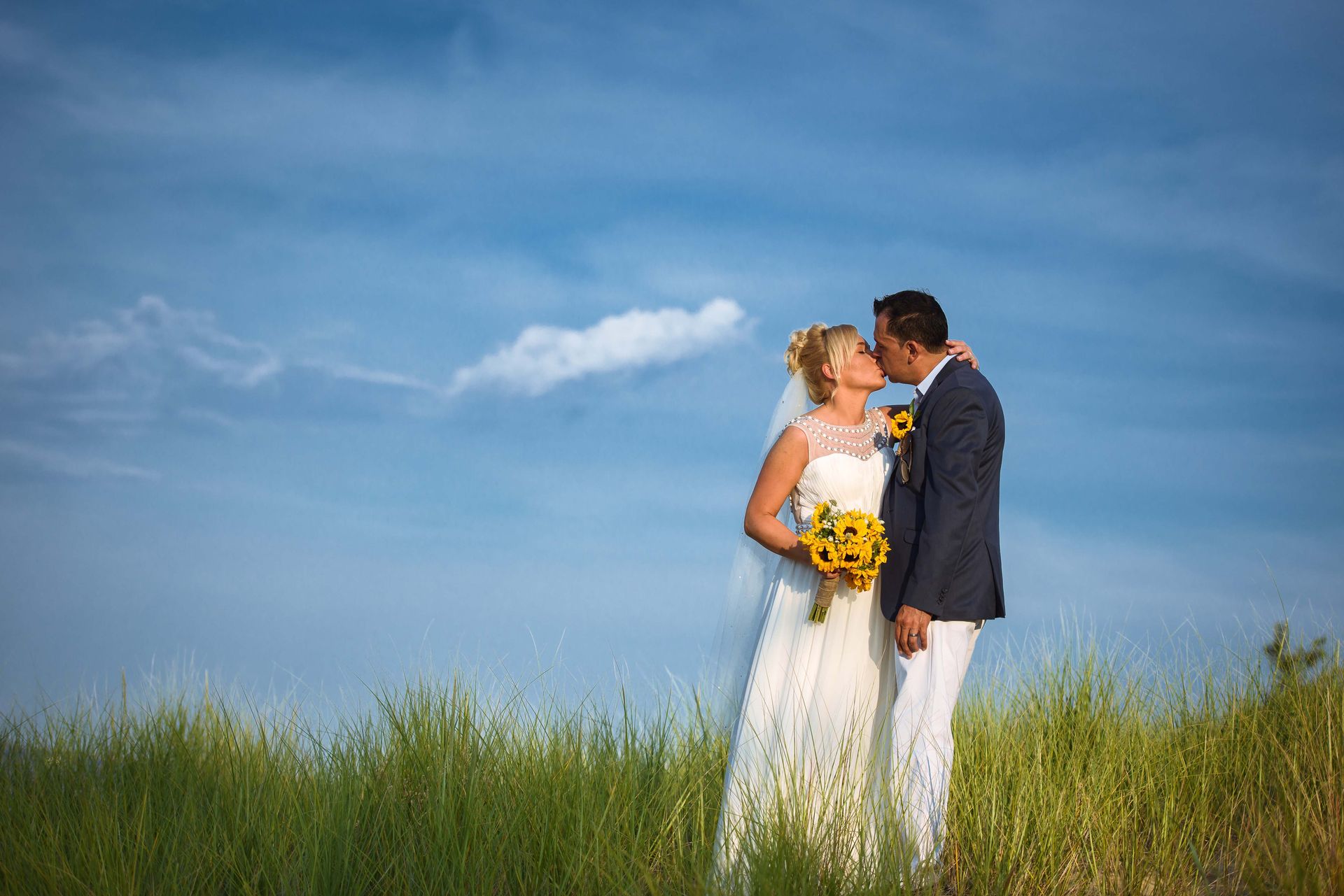 Bride and groom kiss in a field, under a blue sky with a few clouds. Bride holds sunflowers.