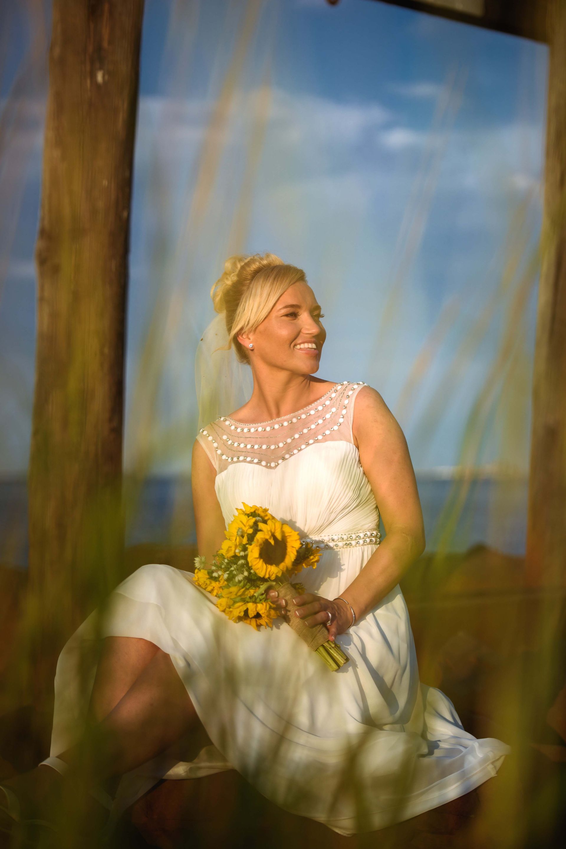 Bride in a white dress with sunflowers, smiling, sitting near water.
