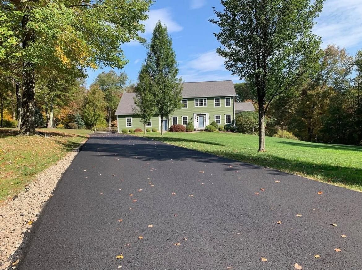 Driveway leading to a beige house with green shutters, trees and lawn in autumn.
