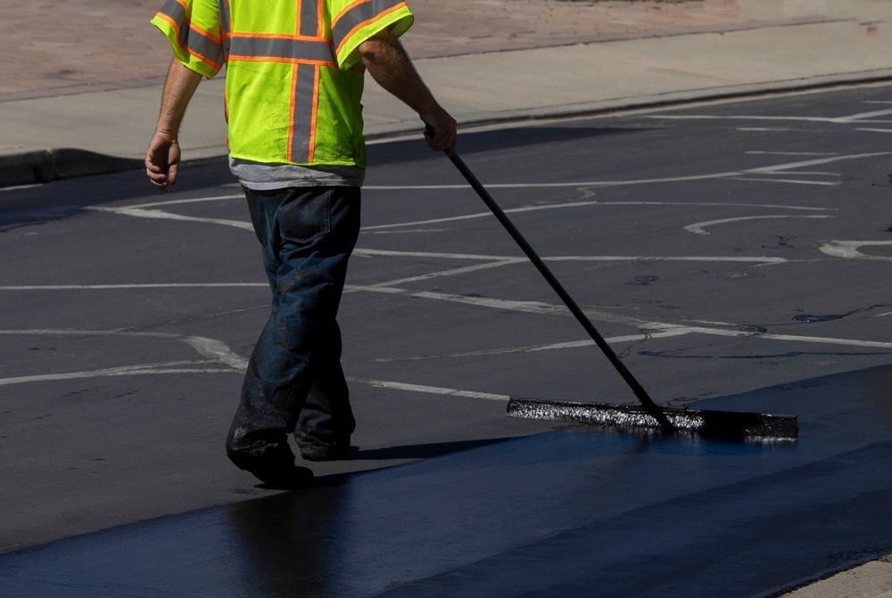Worker in a neon safety vest spreading black asphalt with a long-handled tool on a road.