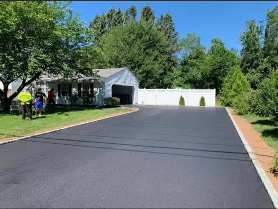 Freshly paved driveway leading to a white garage, with trees and a house on the left