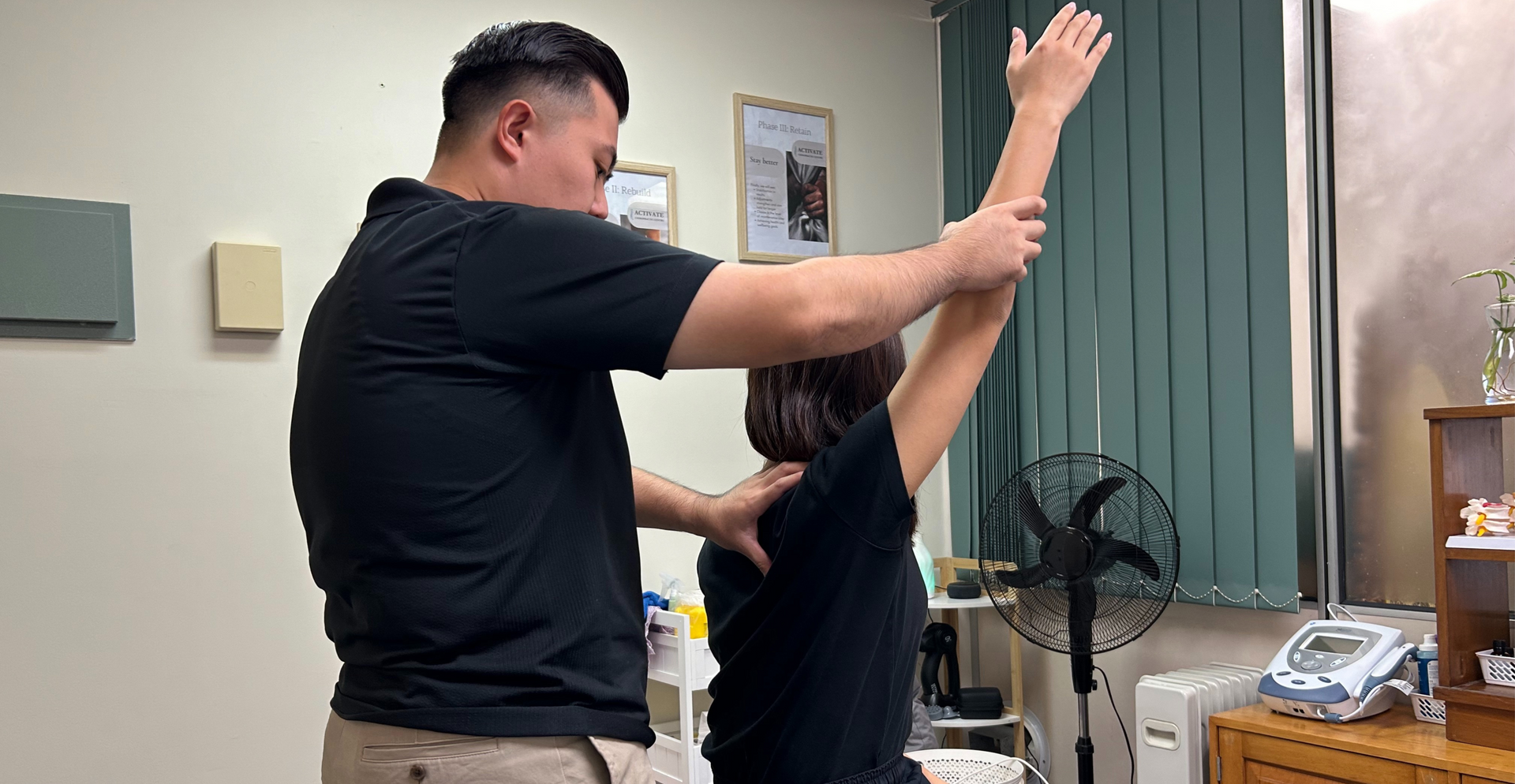 A physical therapist assists a patient in performing an overhead arm stretch in a clinical office setting.