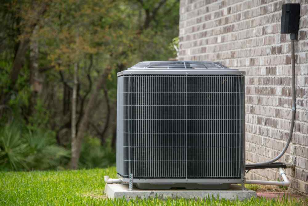 An outdoor HVAC condenser unit sits on a concrete pad next to a brick house with trees in the background.