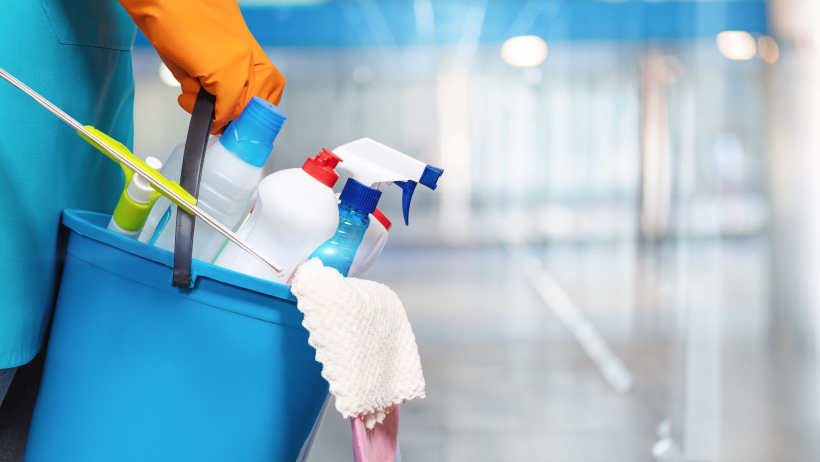 A hand in an orange glove holds a blue bucket filled with various cleaning bottles, a cloth, and a squeegee.