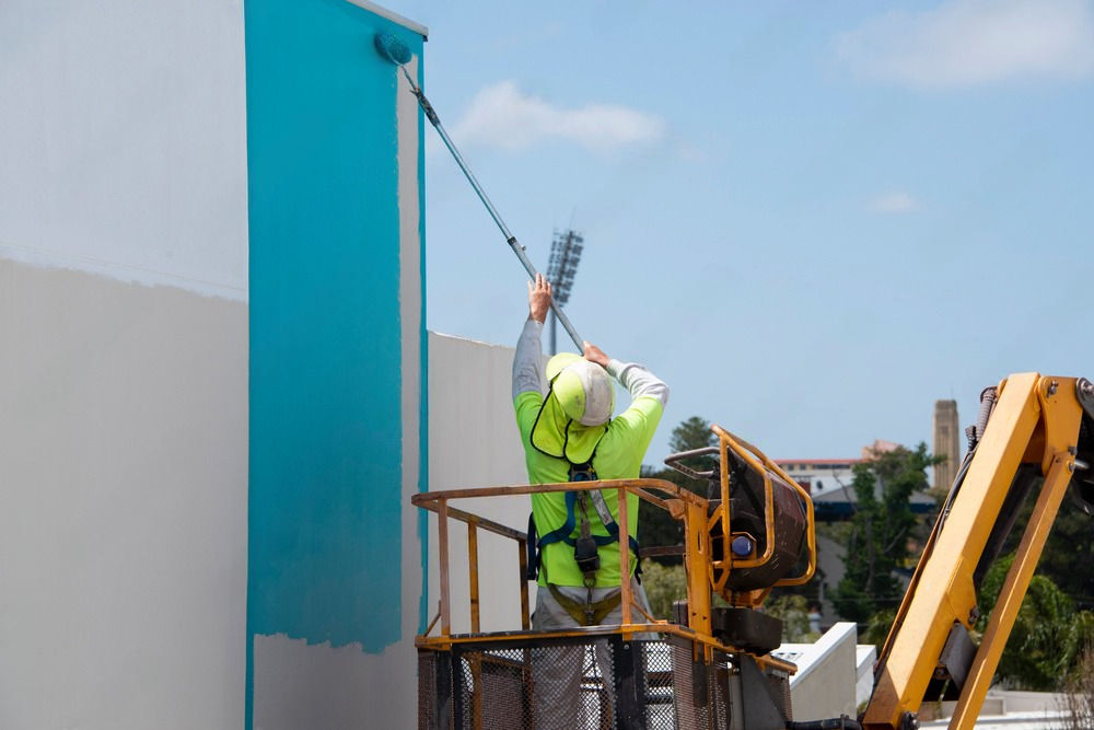 a man is painting a building with a roller while standing on a crane .
