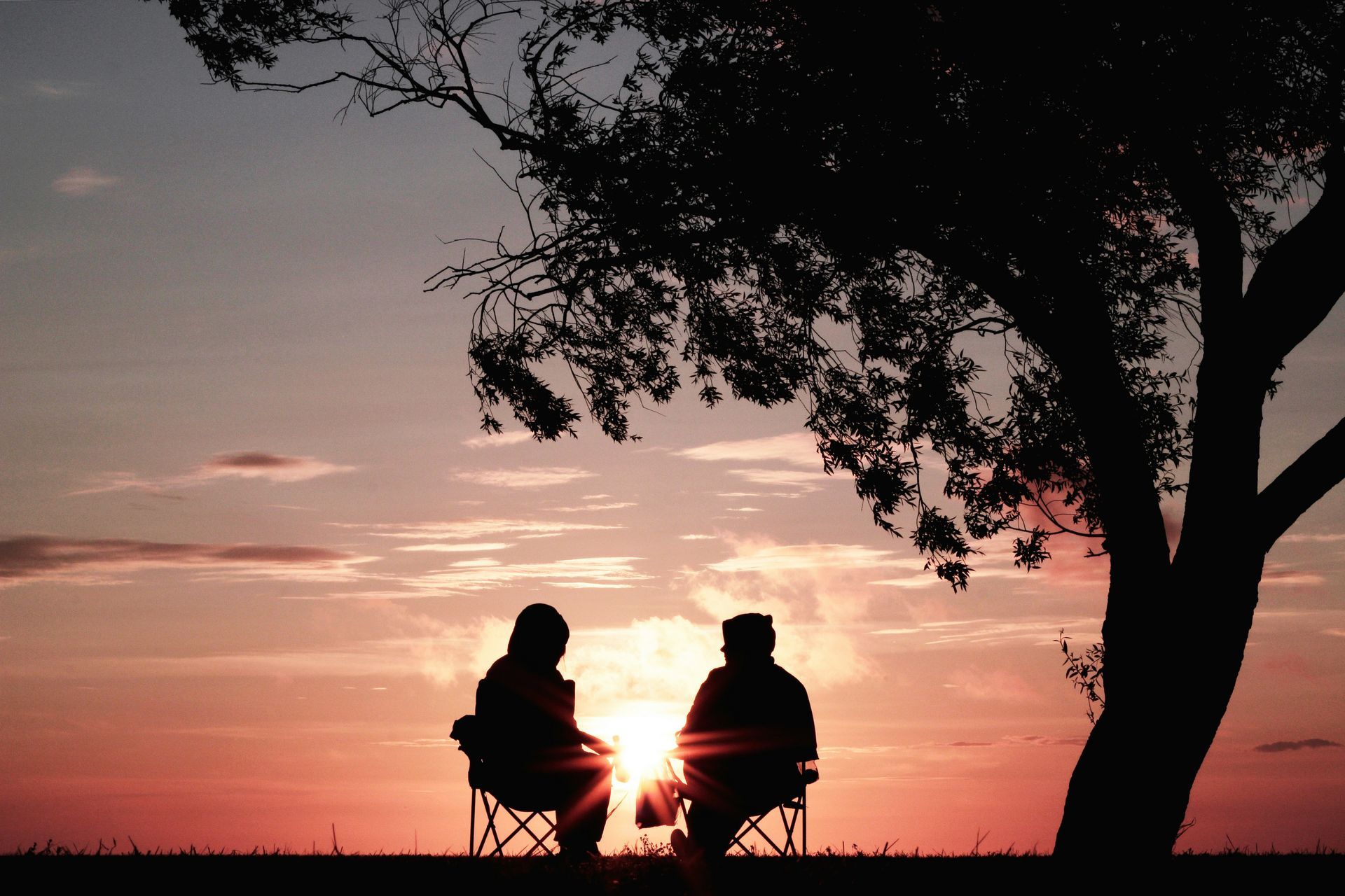 Silhouetted figures seated under a tree at sunset; orange and pink sky.