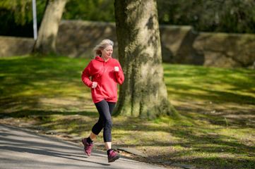 Woman jogs on a path in a park, wearing a red hoodie and black leggings.