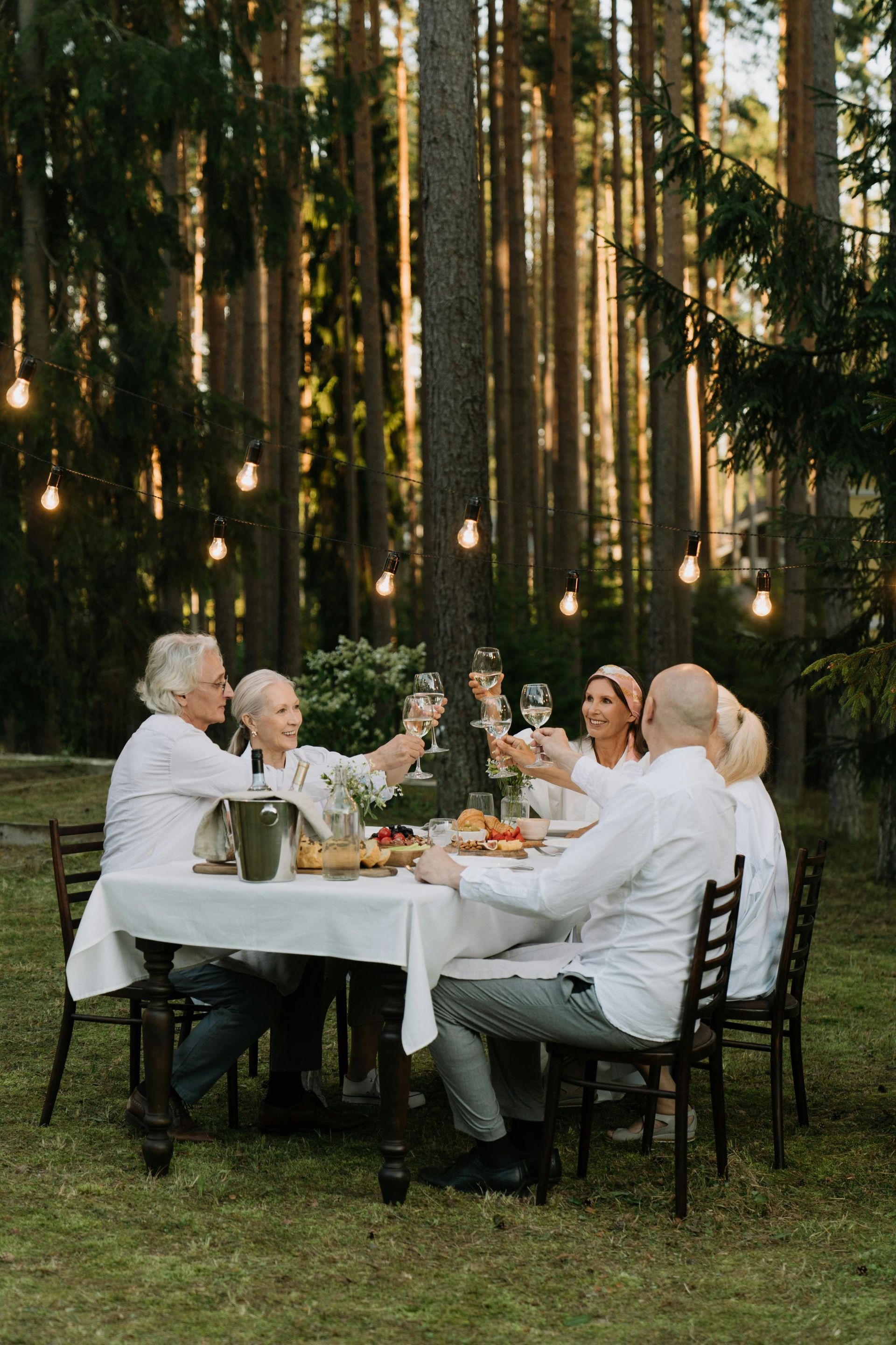 Group of people toasting with wine at a table set outdoors in a forest. String lights hang above.