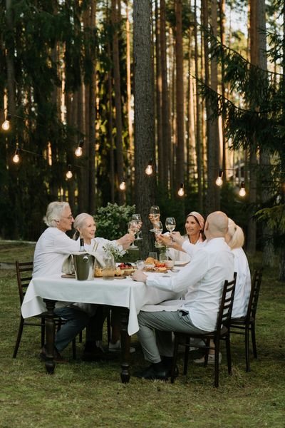 People toasting with wine at a table set for a meal in a forest with string lights.