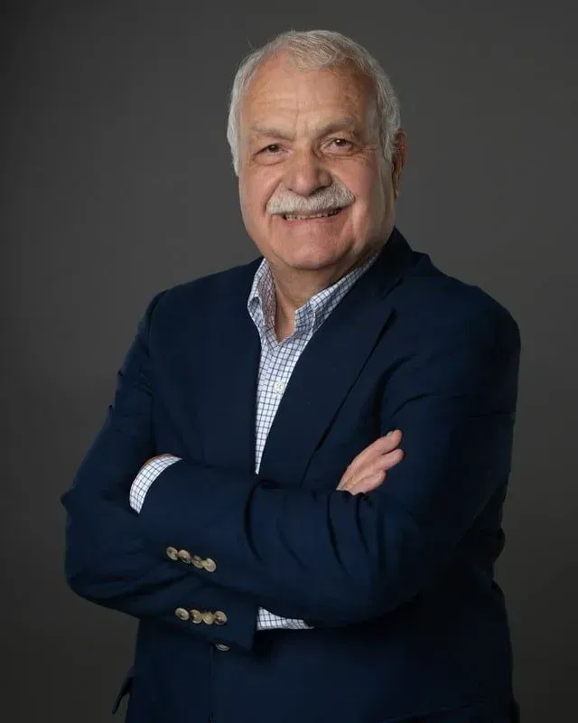 Man with white mustache, arms crossed, wearing a navy blue blazer and patterned shirt. Gray background.