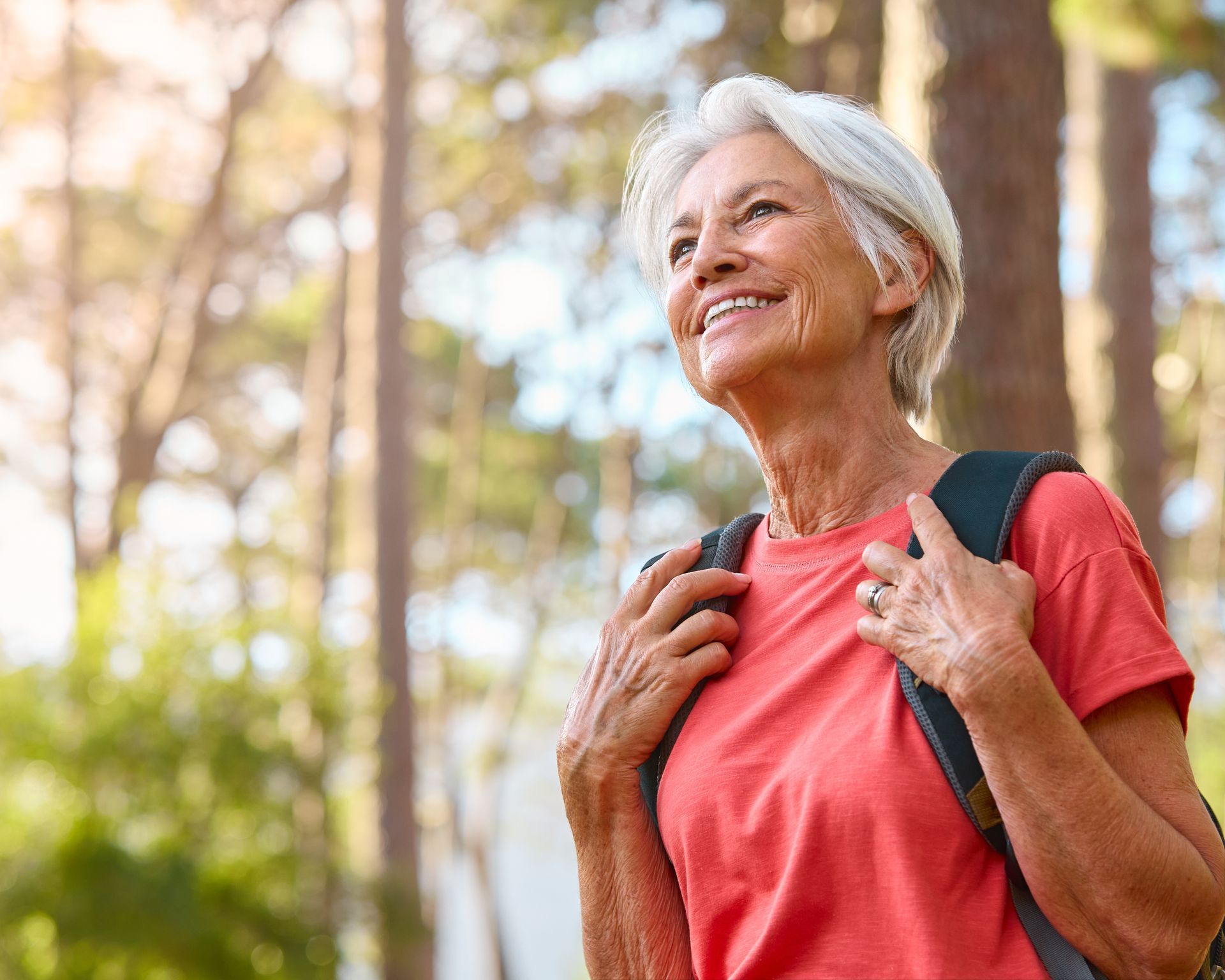 Woman with gray hair smiles while hiking in a forest, wearing a red shirt and backpack.
