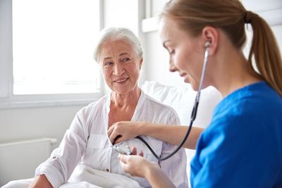 Nurse listening to a patient's chest with a stethoscope. The patient smiles, indoors, bright setting.