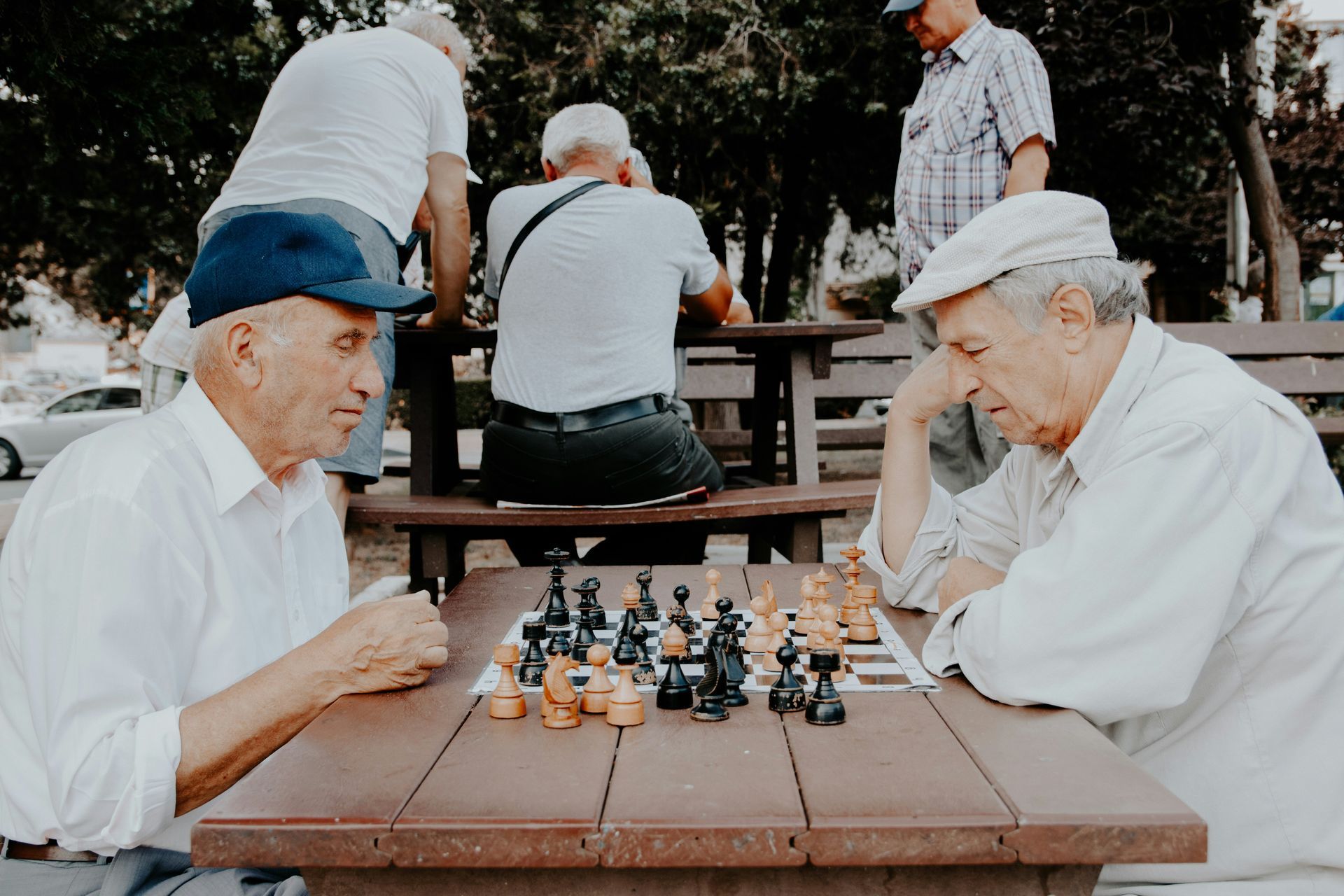 Two men play chess outdoors at a wooden picnic table, watched by other people.
