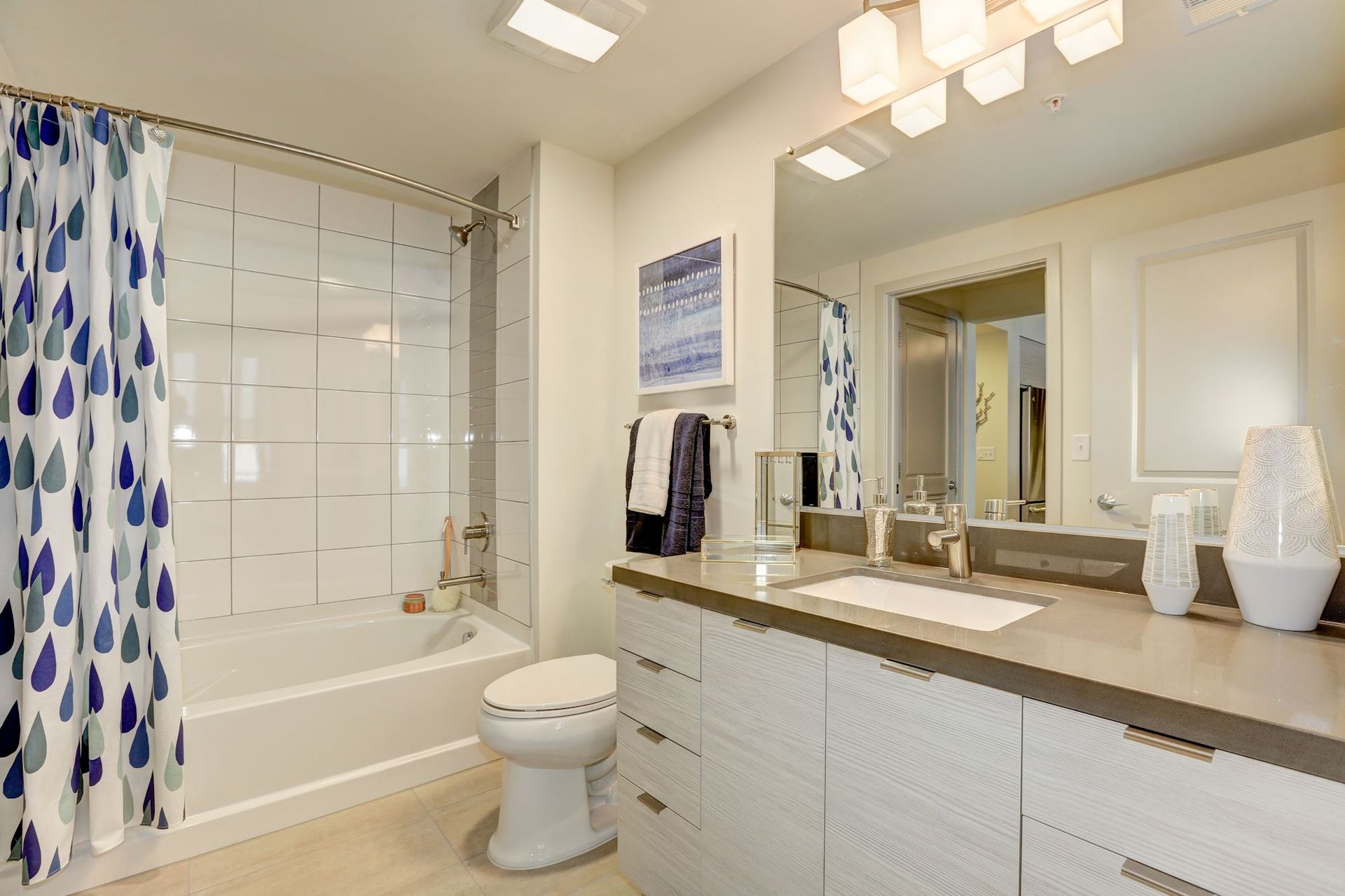 Bright bathroom with white tub, patterned shower curtain, double vanity, and large mirror at Solaire 7077 Woodmont in Bethesda, MD.