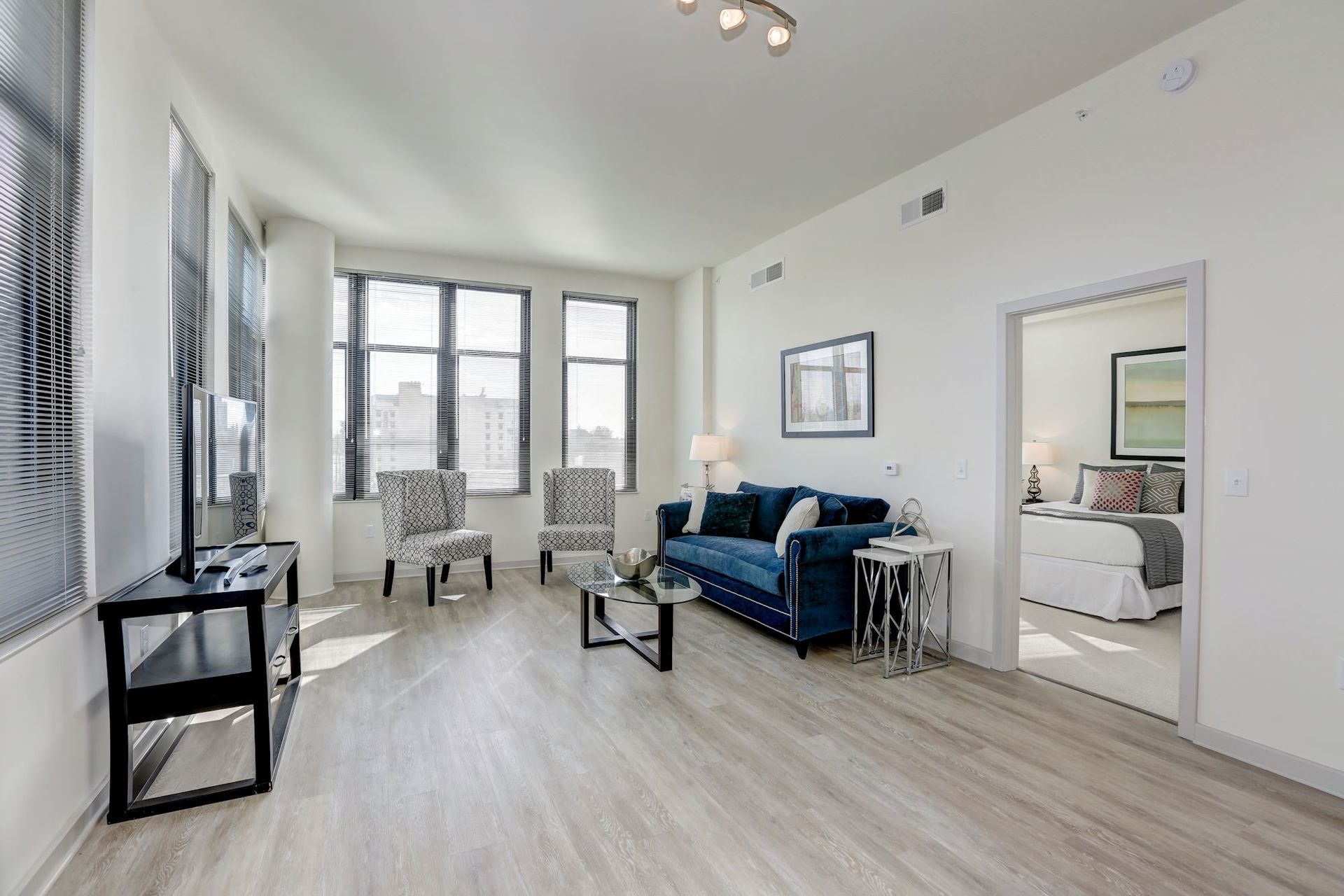 Bright modern living room with blue sofa, chairs, large windows, and an open doorway to a bedroom at Solaire 7077 Woodmont in Bethesda, MD.