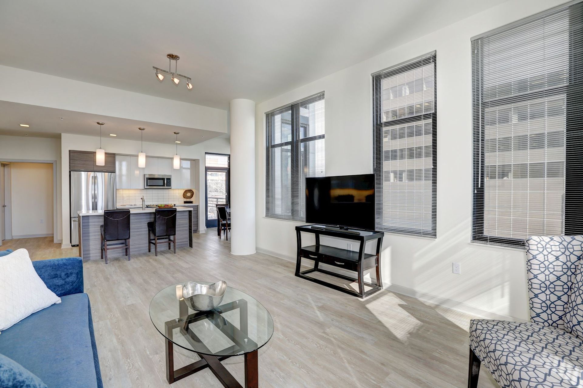 Bright open living room with blue sofa, glass coffee table, TV, and dining area by large windows at Solaire 7077 Woodmont in Bethesda, MD.