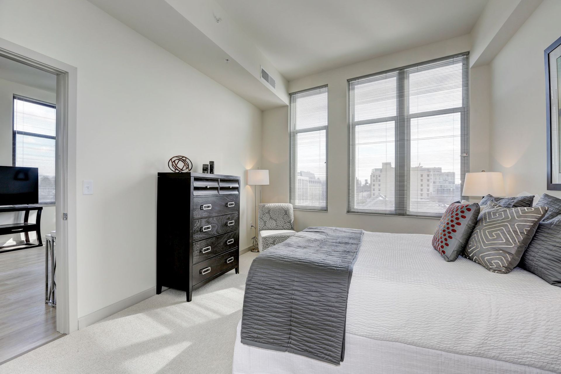 Bright bedroom with a white bed, black dresser, and large windows overlooking a cityscape at Solaire 7077 Woodmont in Bethesda, MD.